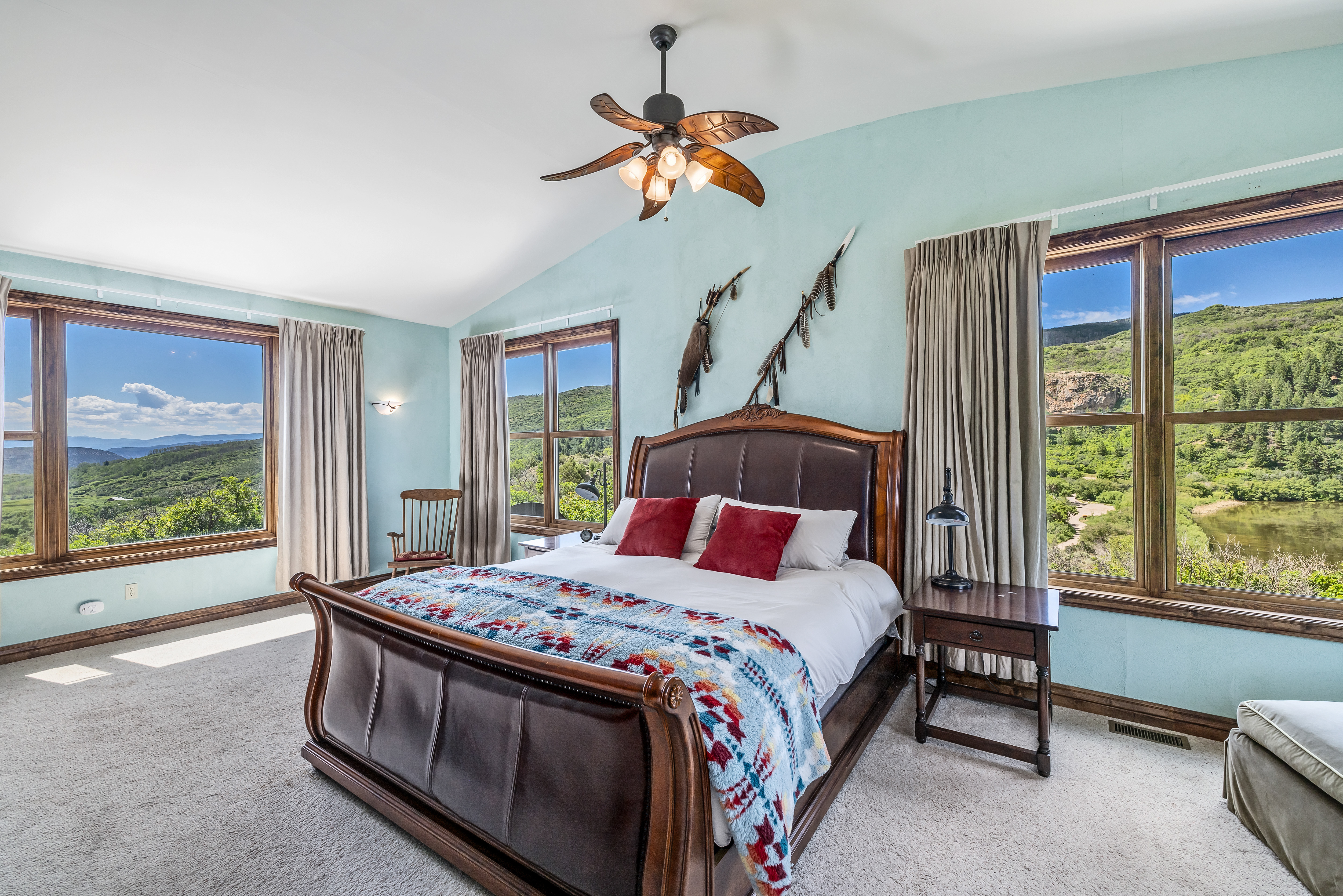 This spacious primary bedroom features a rich, dark wood sleigh bed adorned with a patterned throw blanket and red accent pillows. The room is painted in a soft, light blue, creating a serene atmosphere that is complemented by large windows offering expansive, scenic views of the surrounding hills and water. A ceiling fan with leaf-shaped blades and rustic wall decor add a touch of character to this tranquil, well-lit retreat.