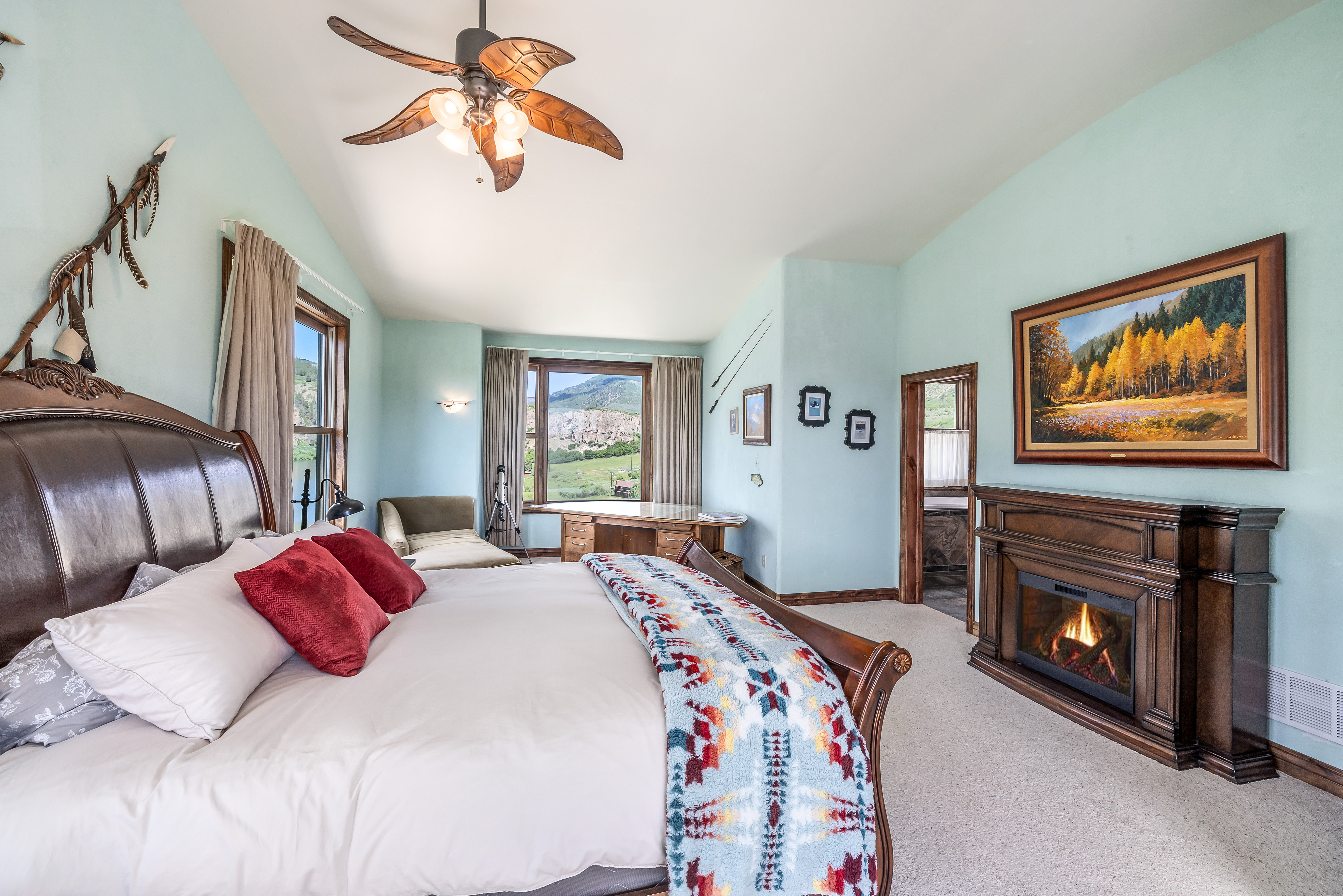 This spacious primary bedroom features a warm, rustic aesthetic with a large wooden bed frame, a cozy fireplace, and a patterned throw blanket. The room is bathed in natural light from large windows that offer a scenic view of the surrounding landscape, complemented by a ceiling fan and soft, light-blue walls. The overall atmosphere is inviting and serene, perfect for a mountain retreat.