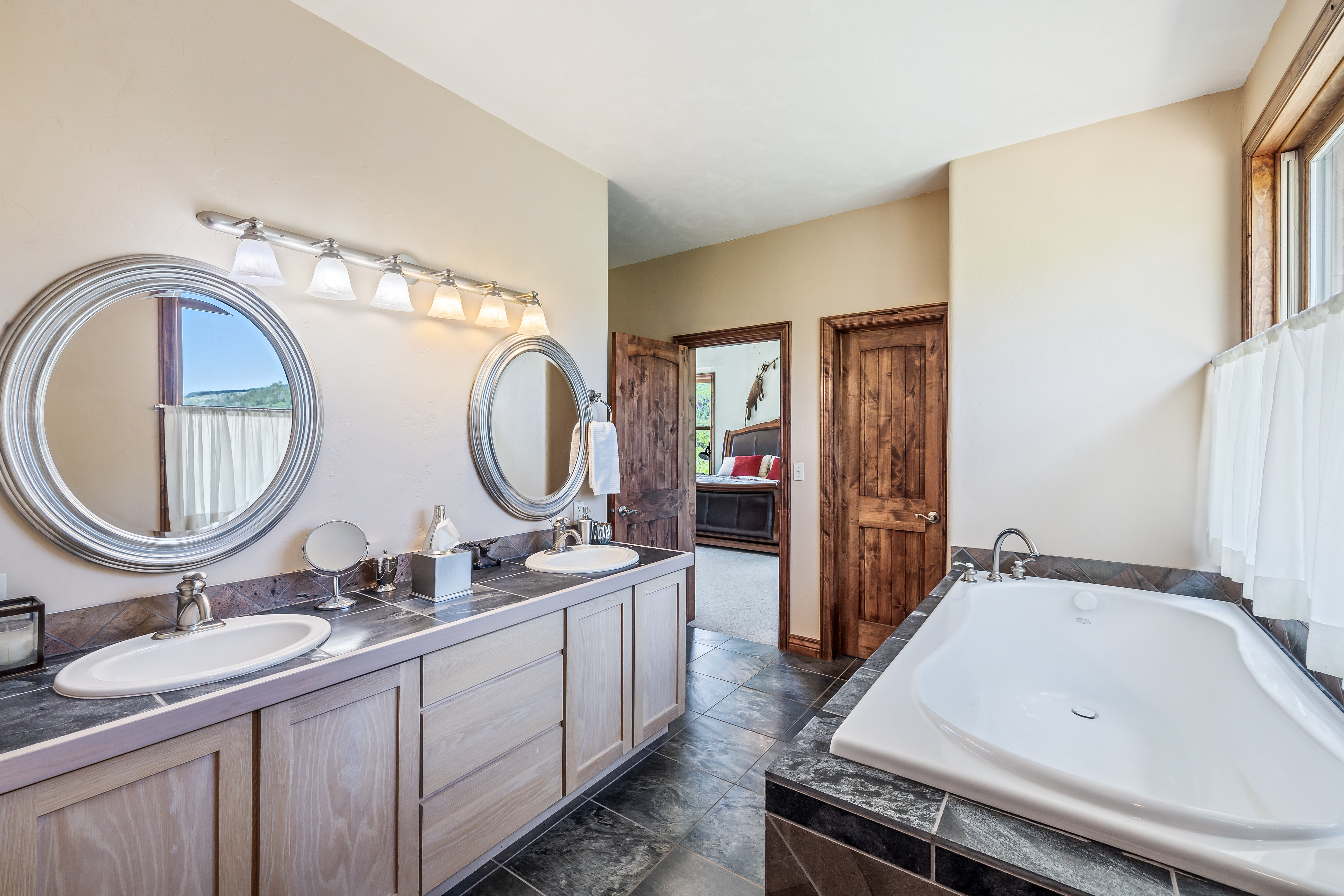 This spacious primary bathroom features a dual-sink vanity with light wood cabinetry and dark stone countertops, complemented by a large soaking tub set in a matching stone surround. The room is illuminated by a multi-light vanity fixture and features elegant round mirrors, with a doorway providing a glimpse into the adjacent bedroom. The overall aesthetic is rustic and refined, with dark slate-style floor tiles grounding the neutral color palette.