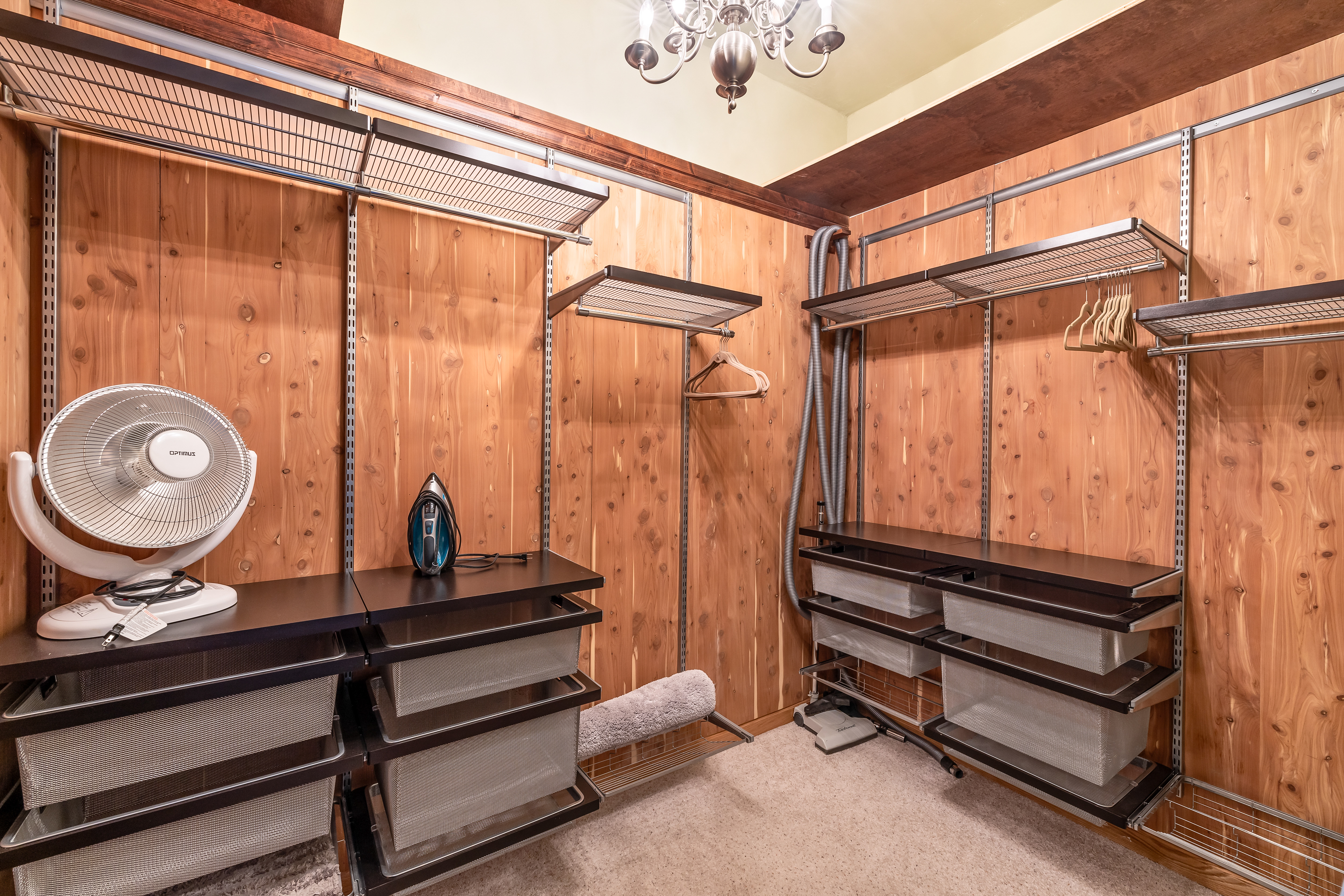 This walk-in closet features warm cedar wood paneling throughout, creating a cozy and aromatic atmosphere. The space is organized with modular wire shelving, pull-out metal storage baskets, and hanging rods, providing ample storage for clothing and accessories. A small desk fan and an iron are placed on the counter, while a central chandelier adds a touch of elegance to the functional design.