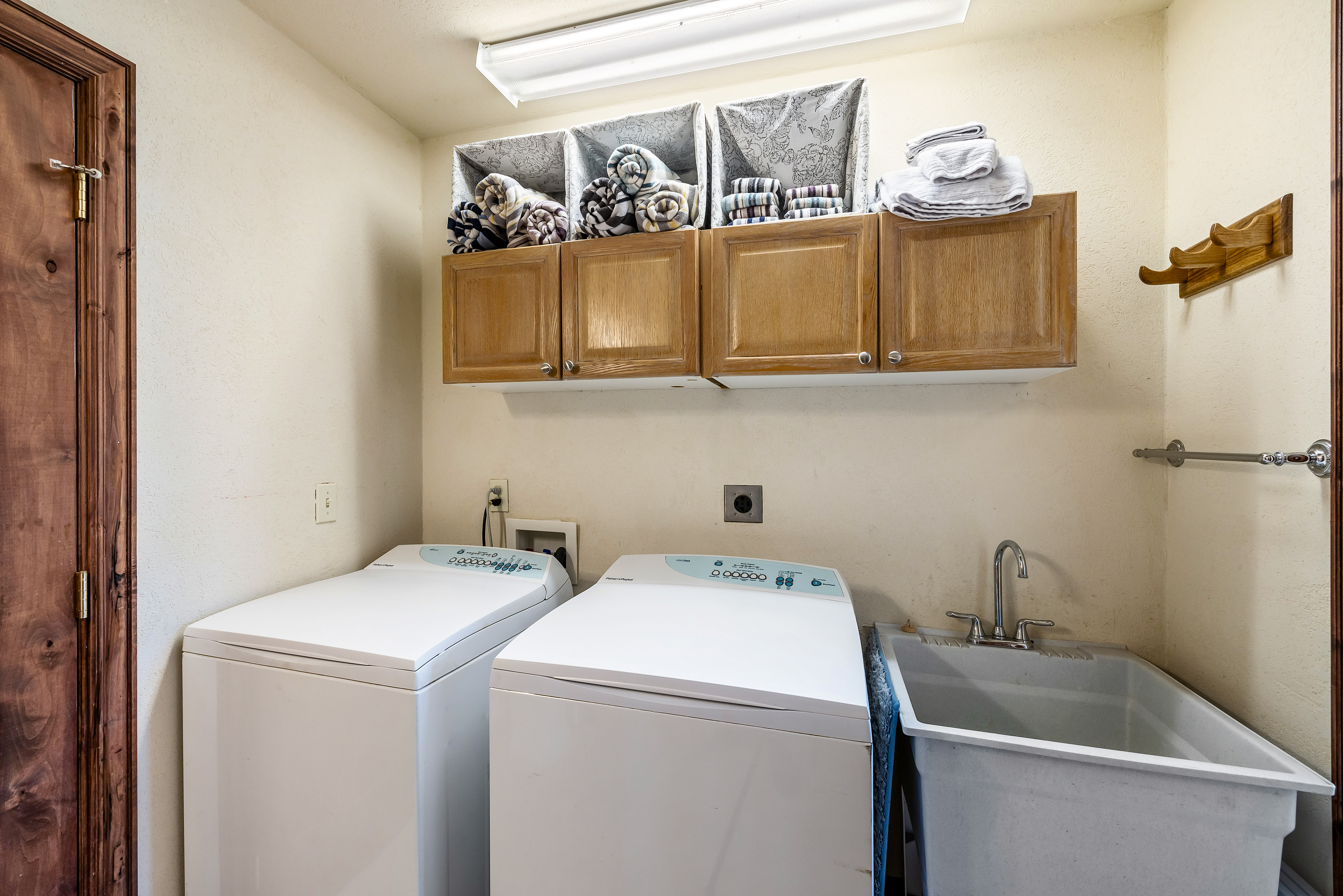 This functional laundry room features a side-by-side washer and dryer set positioned next to a utility sink. Above the appliances, wooden cabinets provide storage, topped with fabric bins and folded linens, while a wooden wall-mounted rack and a towel bar offer additional utility. The room is brightly lit by a ceiling-mounted fluorescent fixture, creating a clean and practical workspace.