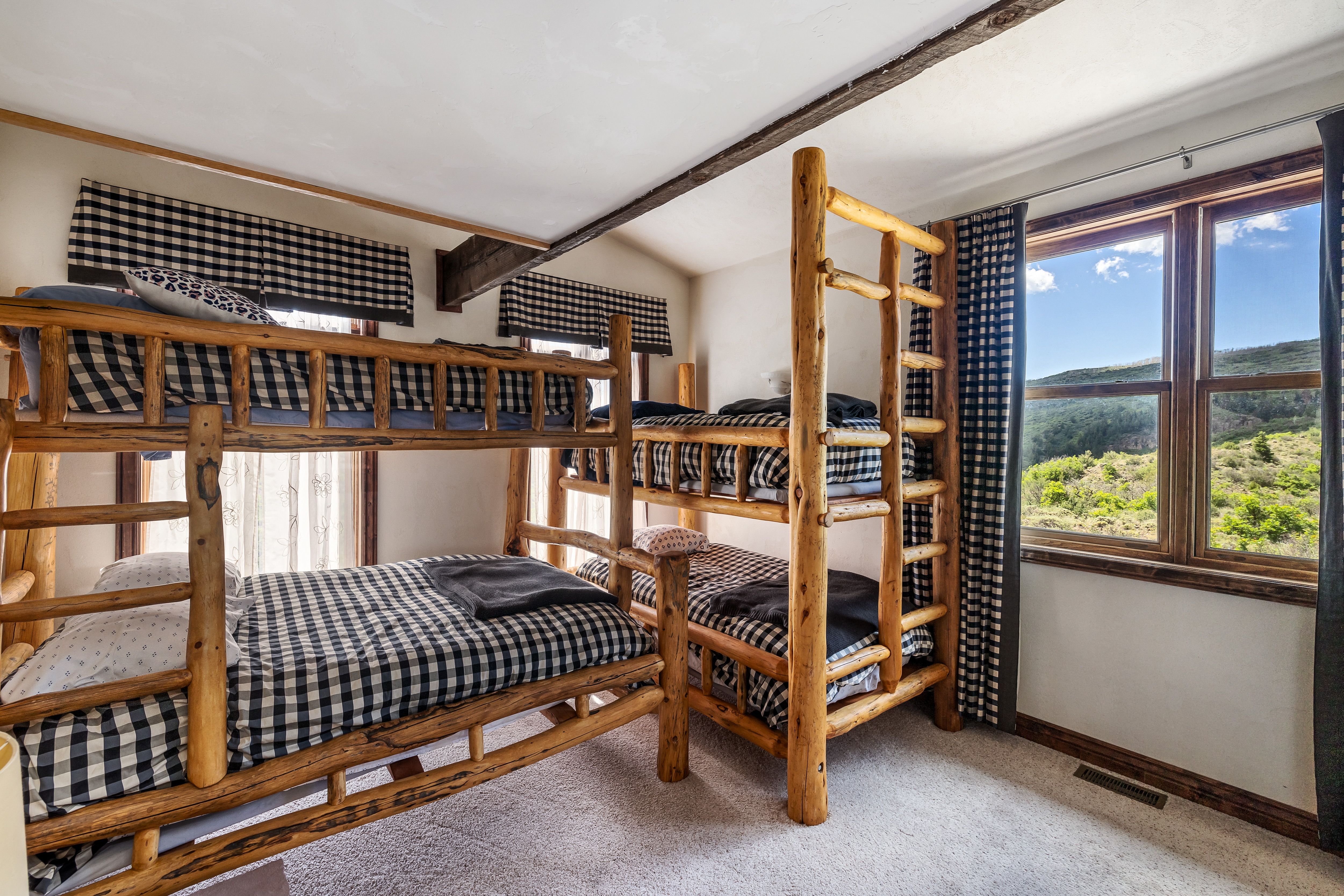 This cozy bedroom features two rustic log-style bunk beds with matching black and white gingham bedding, creating a charming cabin-inspired aesthetic. A large window provides a scenic view of the surrounding hills, while exposed wooden ceiling beams add to the room's mountain lodge character. The space is carpeted and well-lit, offering a comfortable and inviting atmosphere for guests or children.