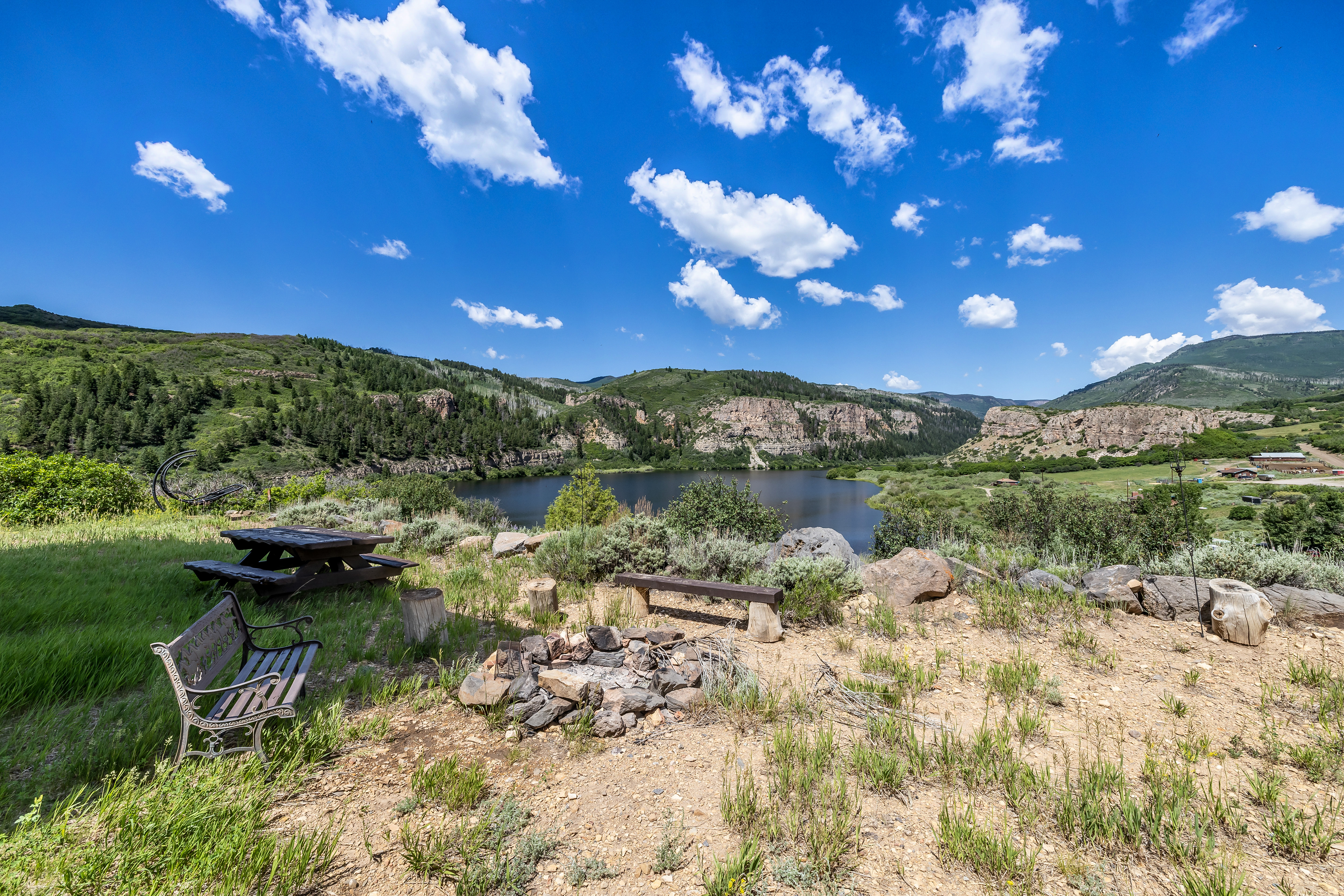 This scenic outdoor space features a rustic fire pit area, a wooden bench, and a picnic table overlooking a serene lake nestled between rolling, tree-covered mountains. The foreground is composed of natural, rugged terrain with native grasses and rocks, creating a peaceful and expansive atmosphere. The wide-angle perspective captures the vastness of the landscape, emphasizing the property's connection to the surrounding wilderness.