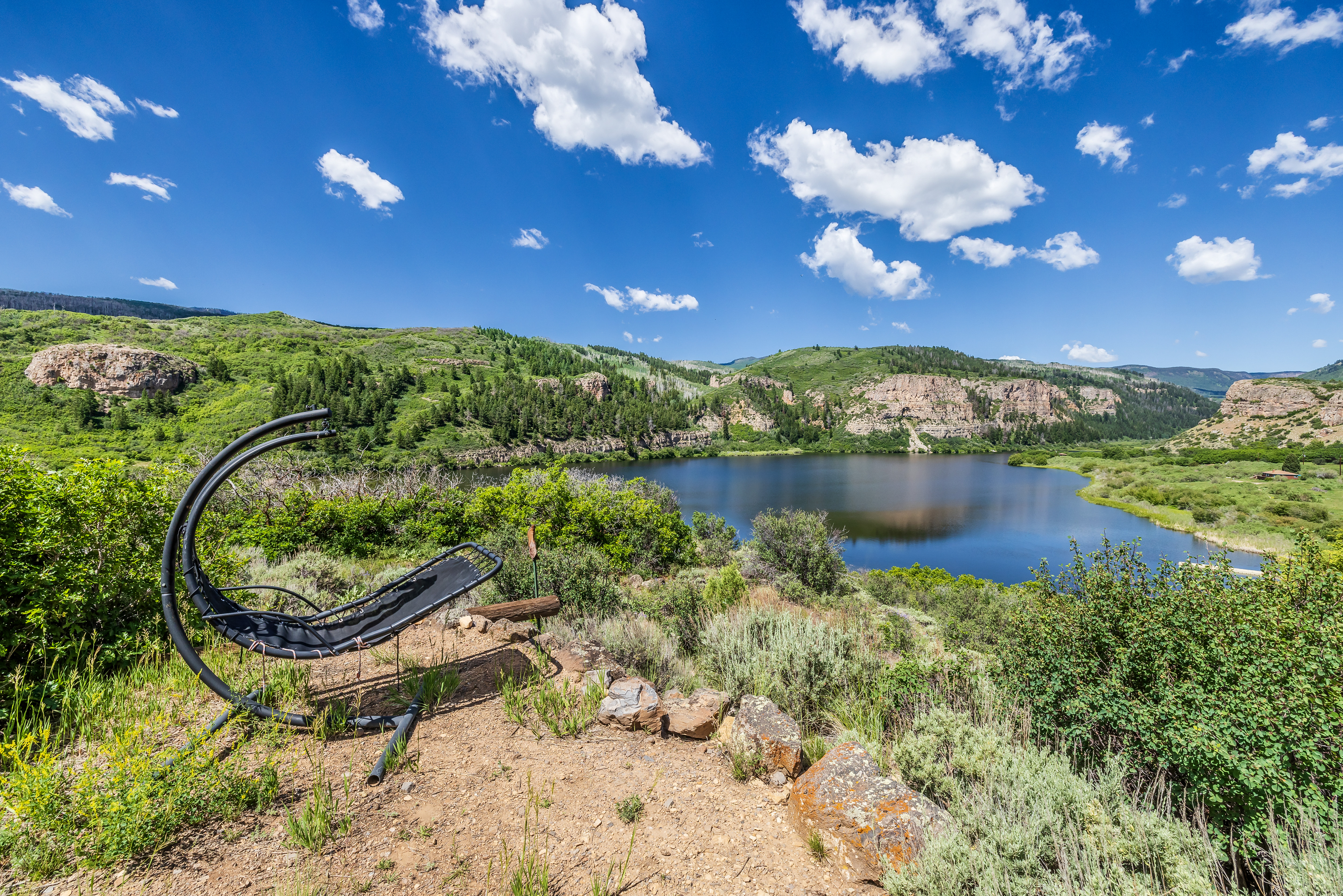 This scenic outdoor space features a modern, curved lounge chair positioned on a dirt overlook, providing a panoramic view of a serene lake nestled between rolling, tree-covered hills. The landscape is characterized by natural, rugged terrain with native shrubs and rock formations, creating a peaceful and secluded atmosphere. The bright blue sky with scattered clouds enhances the expansive, picturesque quality of this property's natural surroundings.