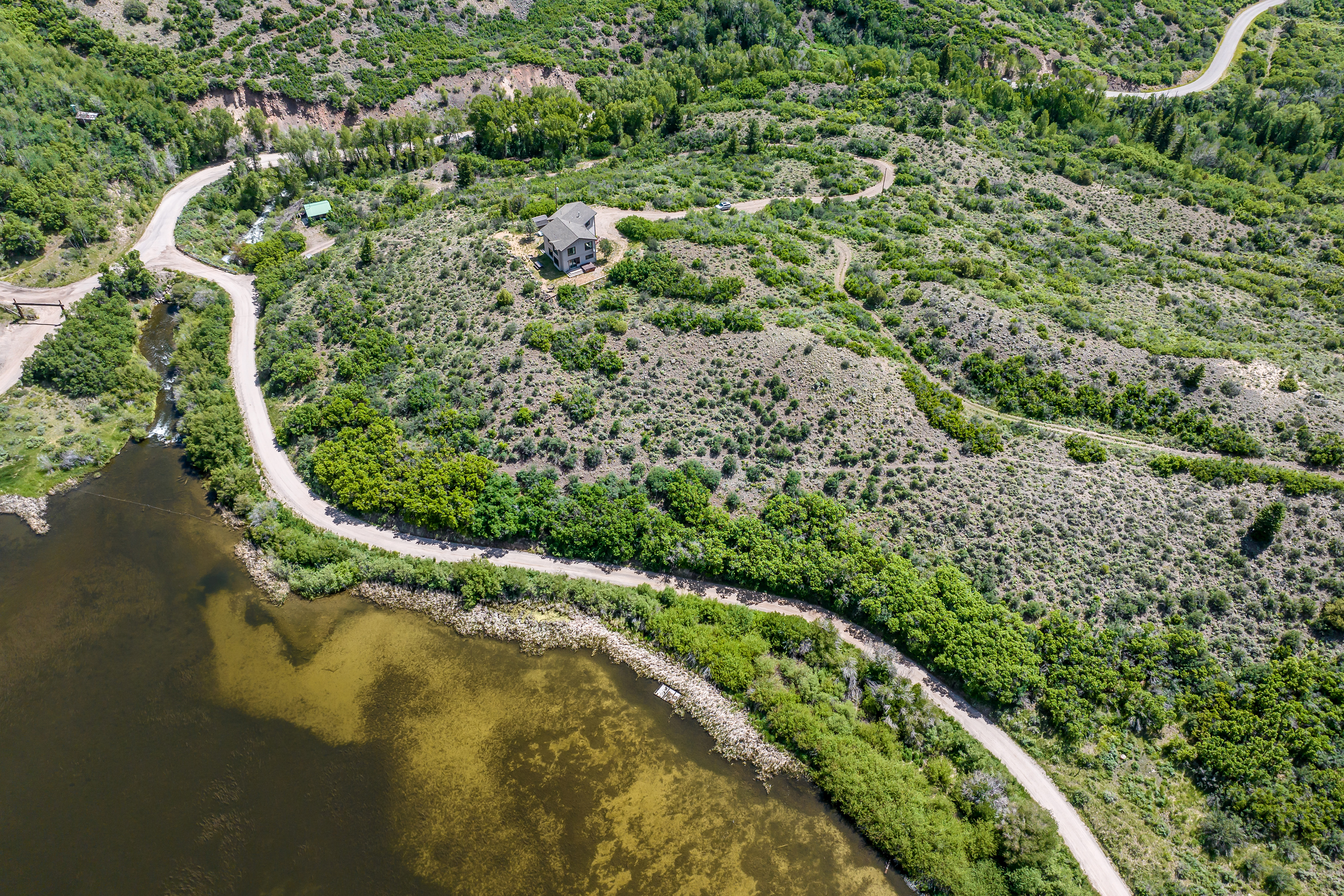 This high-angle aerial shot captures a secluded residential property nestled on a grassy hillside overlooking a large, calm lake. The landscape is characterized by rolling terrain, lush green vegetation, and a winding dirt road that leads up to the house. The perspective emphasizes the property's privacy, natural beauty, and expansive outdoor surroundings.