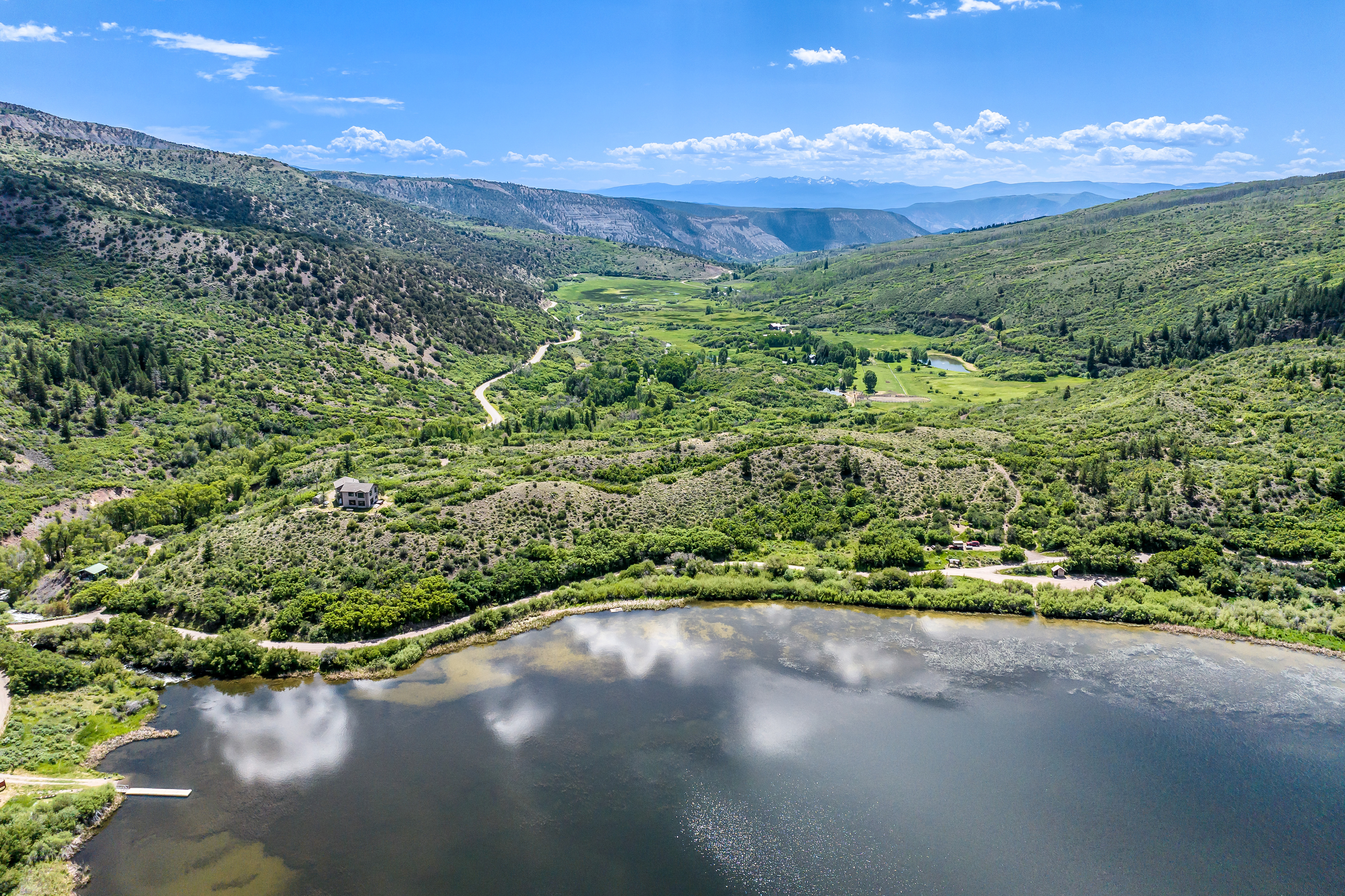 This stunning aerial view captures a serene mountain landscape featuring a large, reflective lake in the foreground and a sprawling valley surrounded by lush, green hills. A single residential home is prominently situated on a hillside overlooking the water, emphasizing the property's secluded and picturesque location. The expansive blue sky and natural terrain create a peaceful, high-end atmosphere ideal for a luxury retreat.