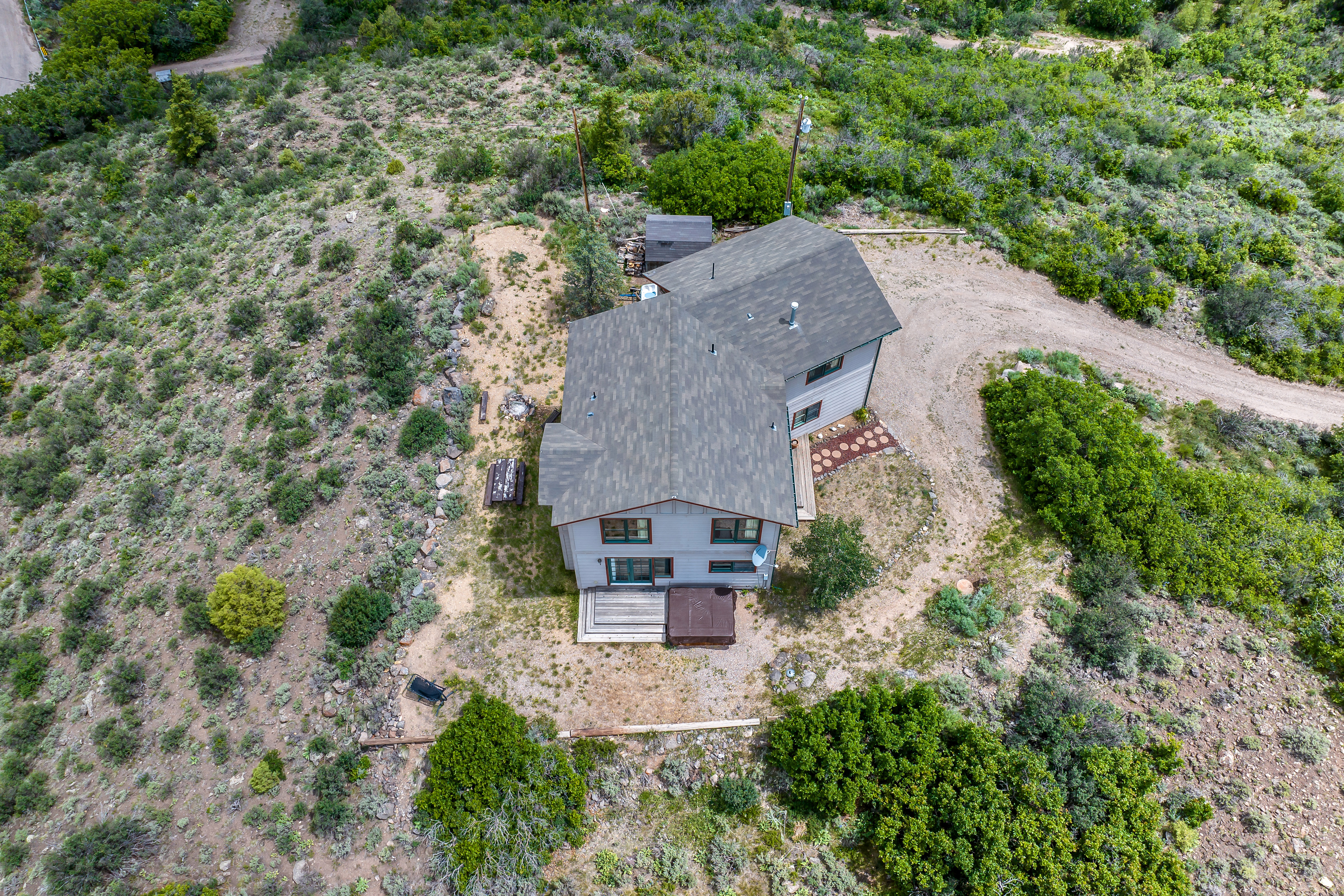 This high-angle aerial view captures a multi-level residential home nestled within a rugged, natural landscape of scrub brush and trees. The property features a shingled roof, a rear deck with a hot tub, and a winding dirt driveway leading up to the structure. The setting offers a secluded, rustic atmosphere, emphasizing the home's integration into the surrounding hillside terrain.