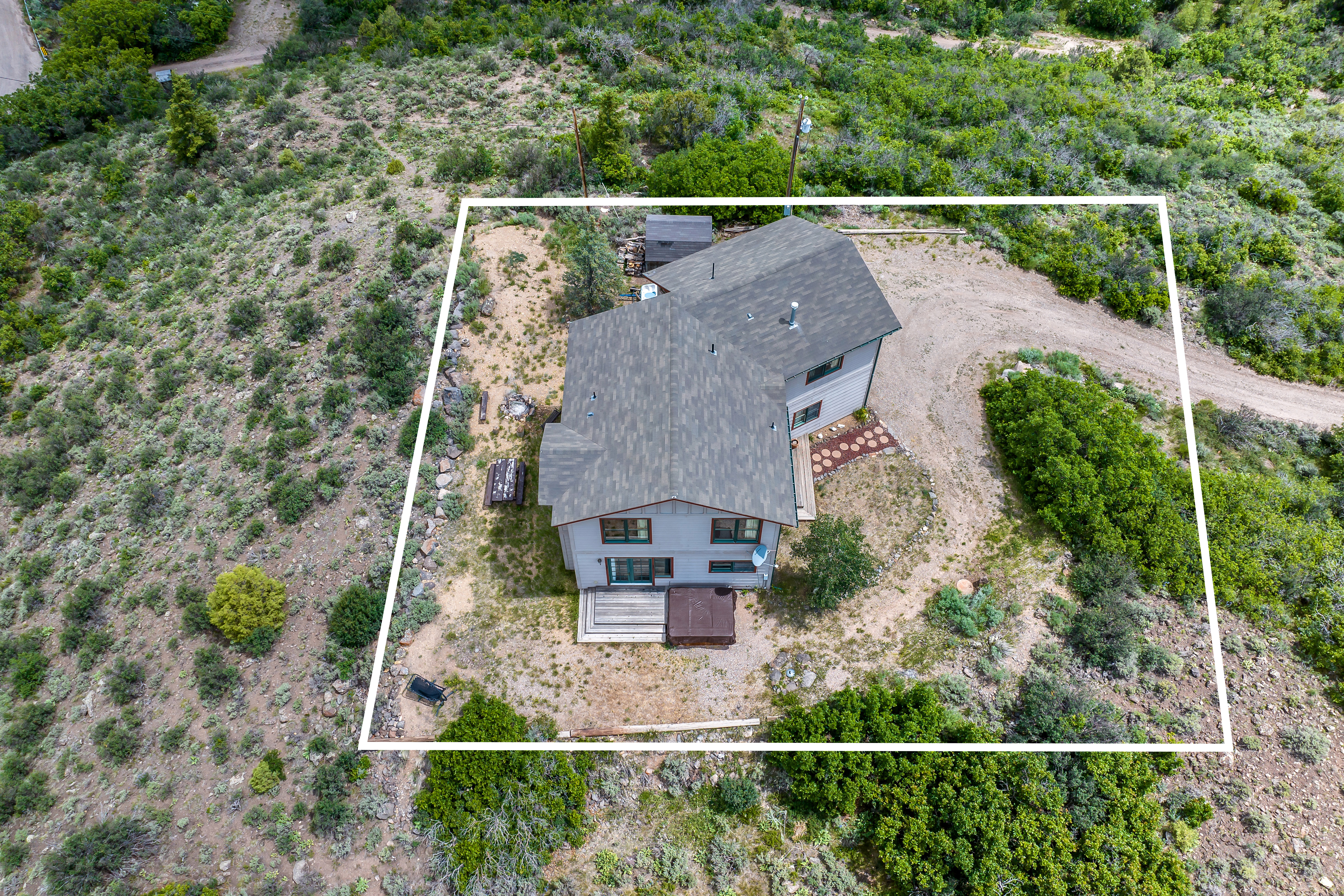 This high-angle aerial view captures a detached single-family home nestled in a rugged, natural landscape. The property features a multi-level roofline, a wooden deck with a hot tub, and a gravel driveway, all framed by a white border overlay. The surrounding terrain is characterized by native brush and trees, emphasizing a secluded, rural setting.