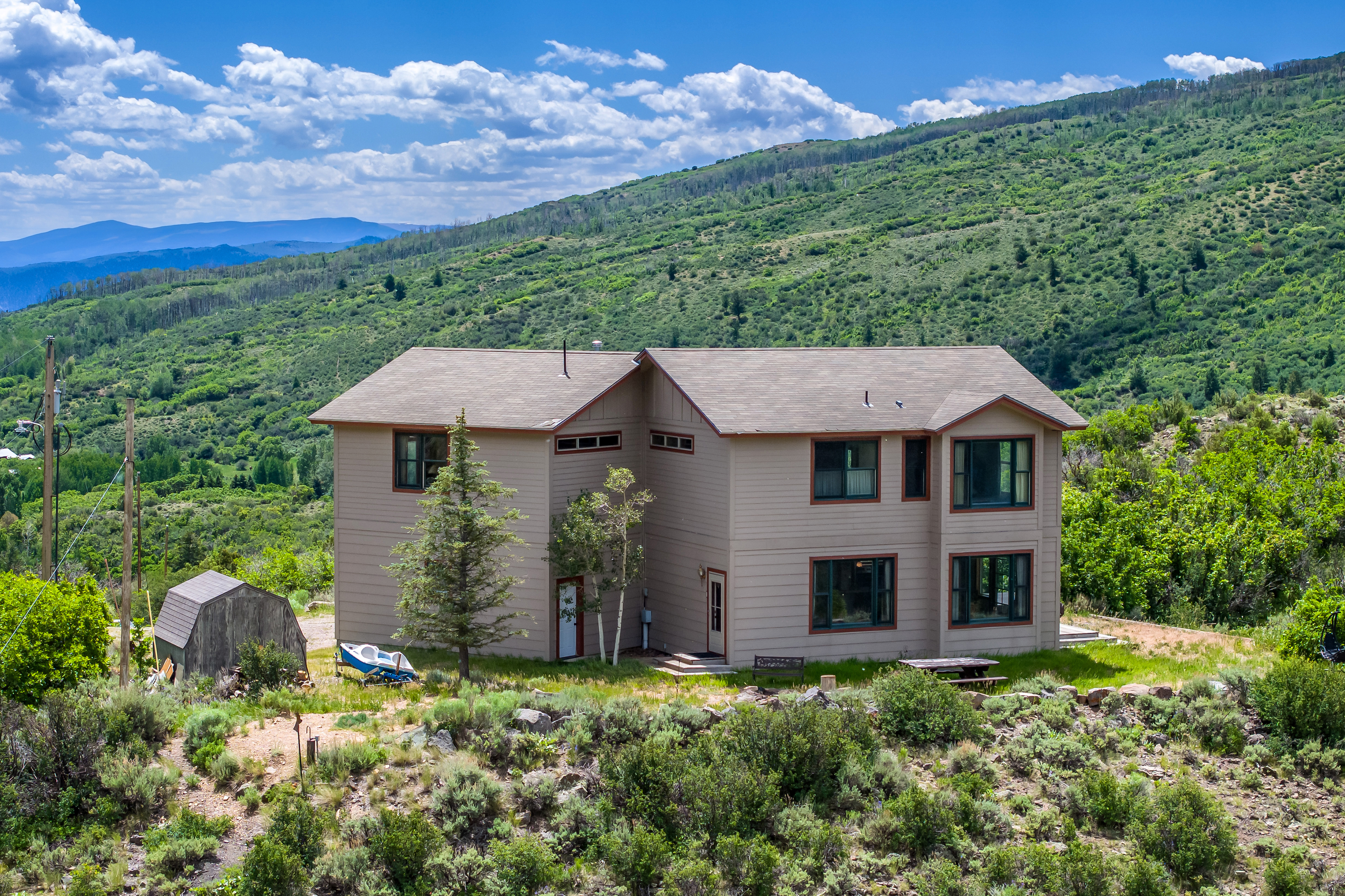 This elevated exterior view captures a two-story, tan-sided home nestled against a lush, green mountain hillside. The property features a simple, functional design with multiple windows, a small shed to the left, and a picnic table on the lawn, evoking a peaceful, secluded mountain retreat atmosphere. The wide-angle perspective emphasizes the home's integration into the expansive, natural landscape under a bright, cloudy sky.