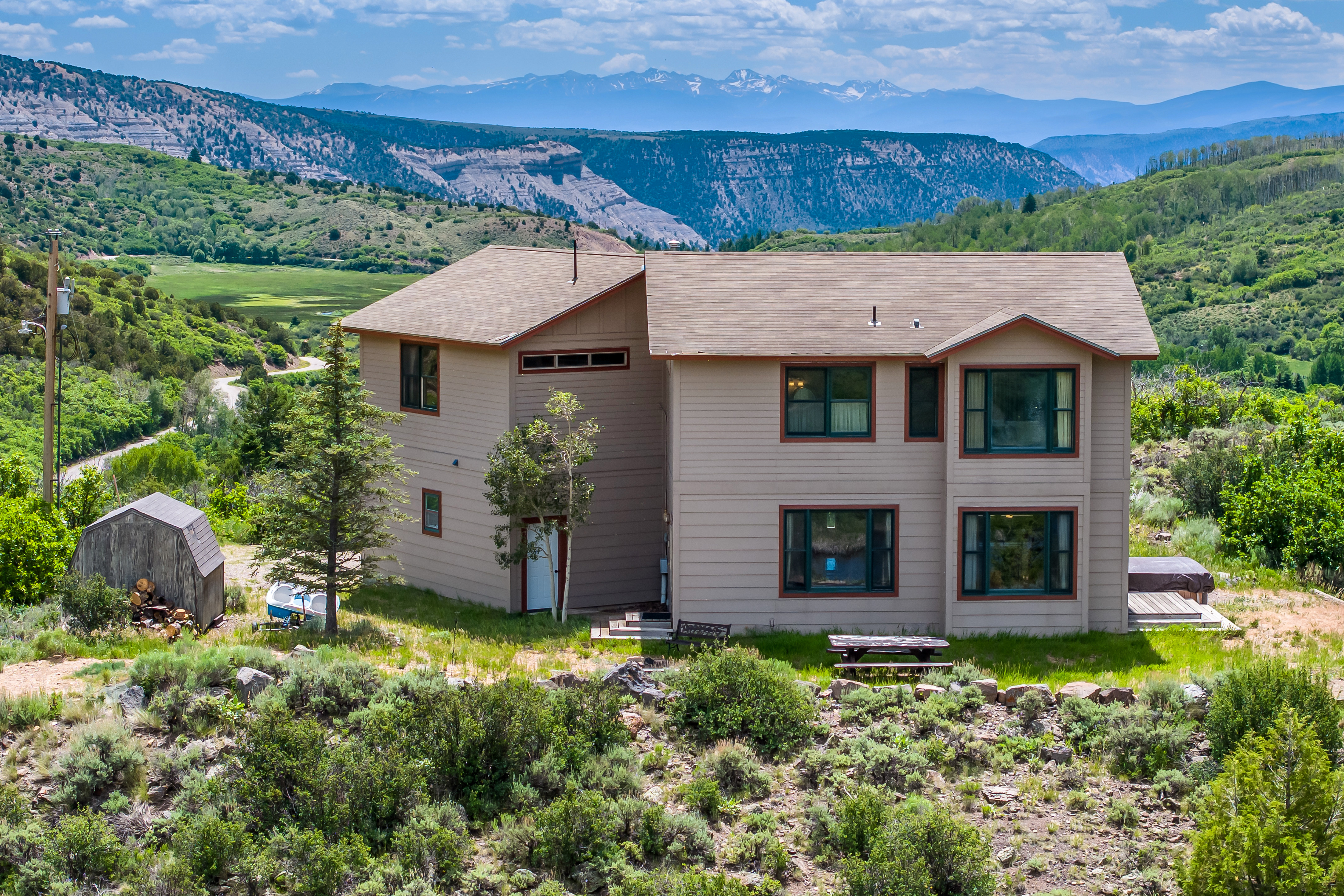 This elevated rear view captures a two-story, light-brown suburban home set against a breathtaking backdrop of rolling green hills and snow-capped mountains. The property features a simple, functional design with large windows, a small wooden deck with a hot tub, and a picnic table in the yard. The surrounding landscape is rugged and natural, emphasizing a peaceful, secluded mountain lifestyle.