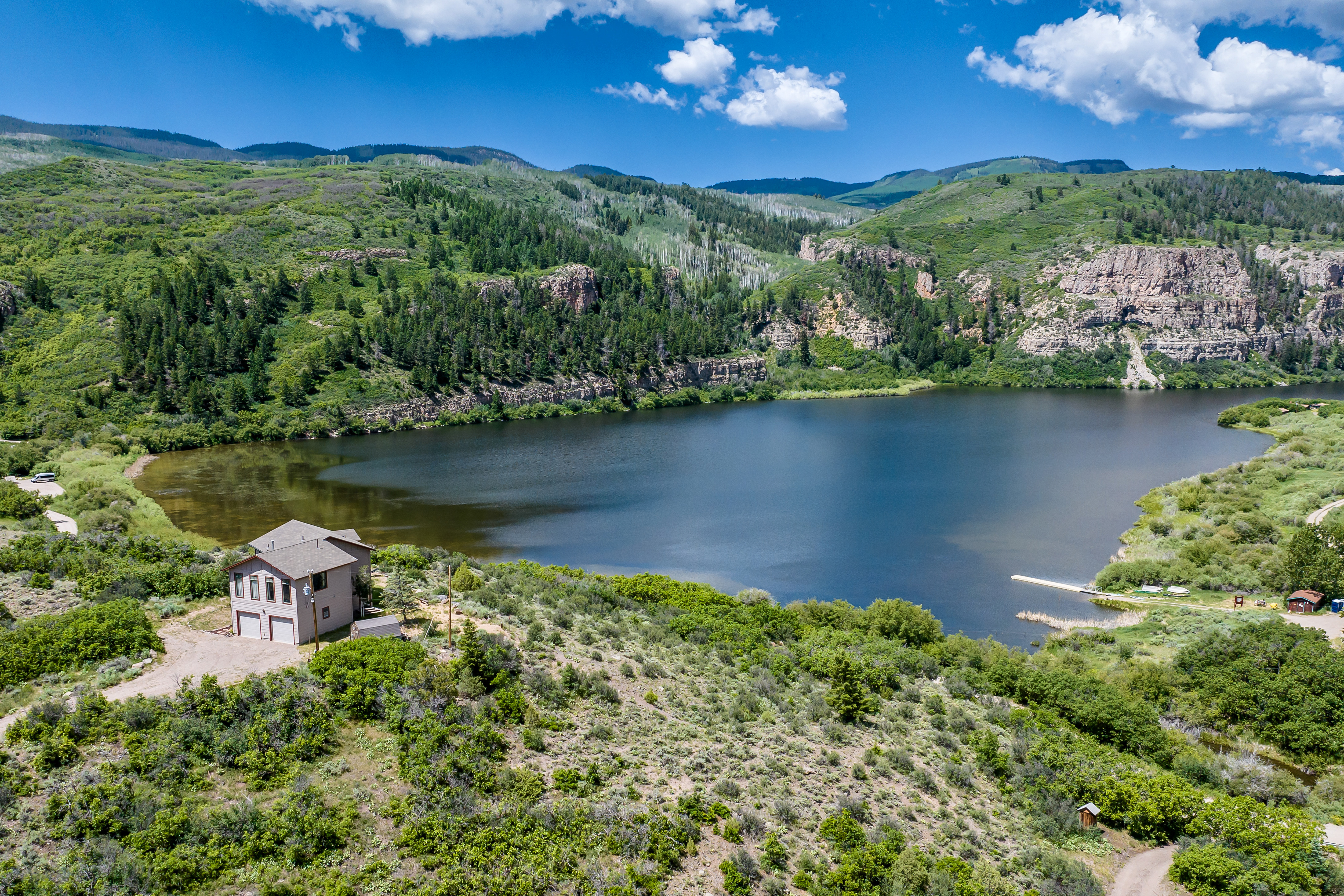 This stunning aerial perspective captures a secluded multi-story home nestled on a hillside overlooking a serene, expansive lake surrounded by lush green mountains and rocky cliffs. The property features a natural, rugged landscape with a private driveway and a small dock extending into the water, emphasizing a peaceful, nature-focused lifestyle. The bright, clear sky and vibrant greenery create an inviting and picturesque setting for a mountain retreat.