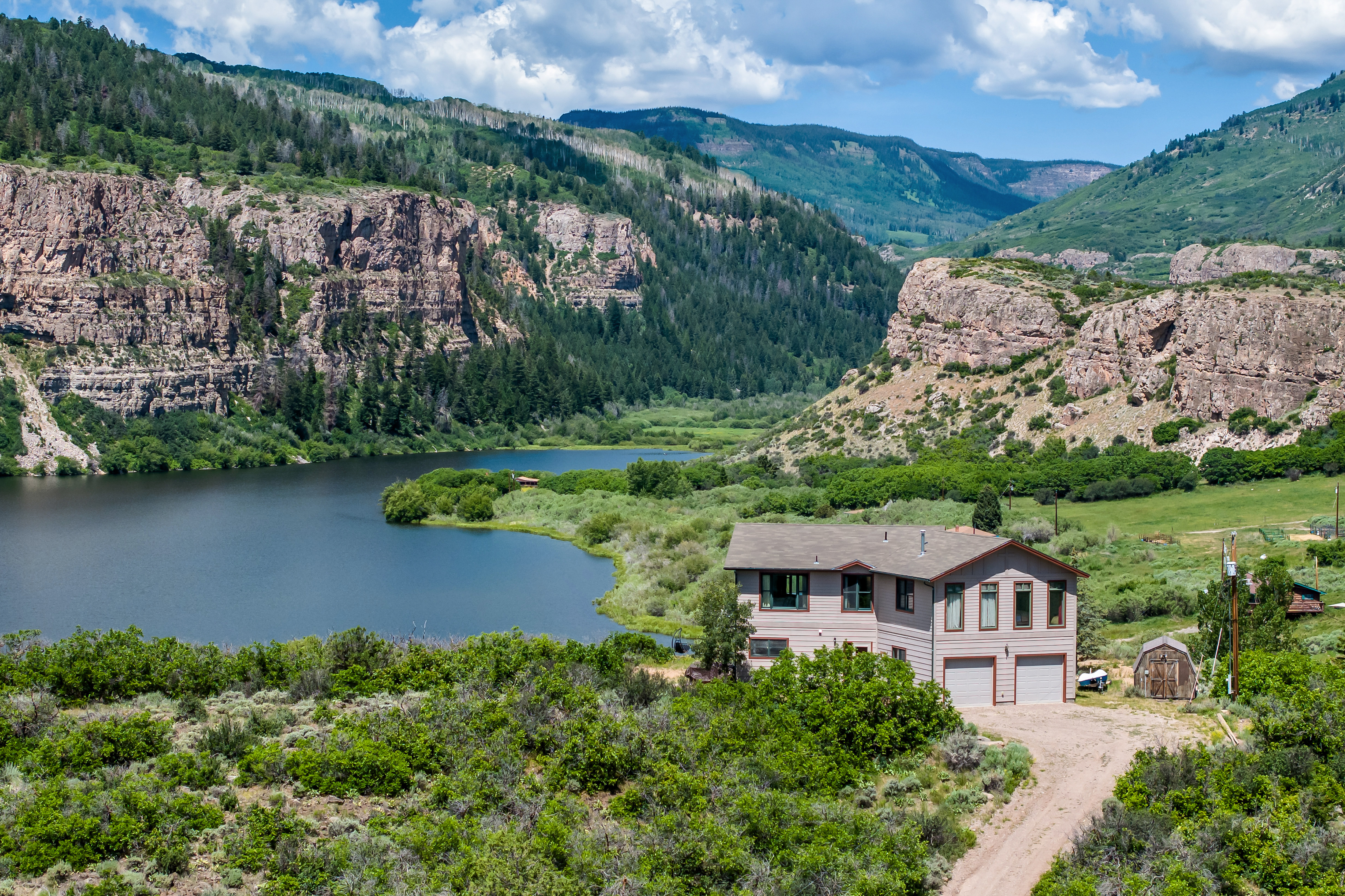 This elevated aerial perspective showcases a multi-level residential home nestled in a stunning, rugged mountain landscape. The property features a two-car garage and is situated directly adjacent to a serene, expansive lake surrounded by lush greenery and dramatic rock cliffs. The overall impression is one of peaceful seclusion and natural beauty, highlighting the home's prime location in a scenic wilderness setting.