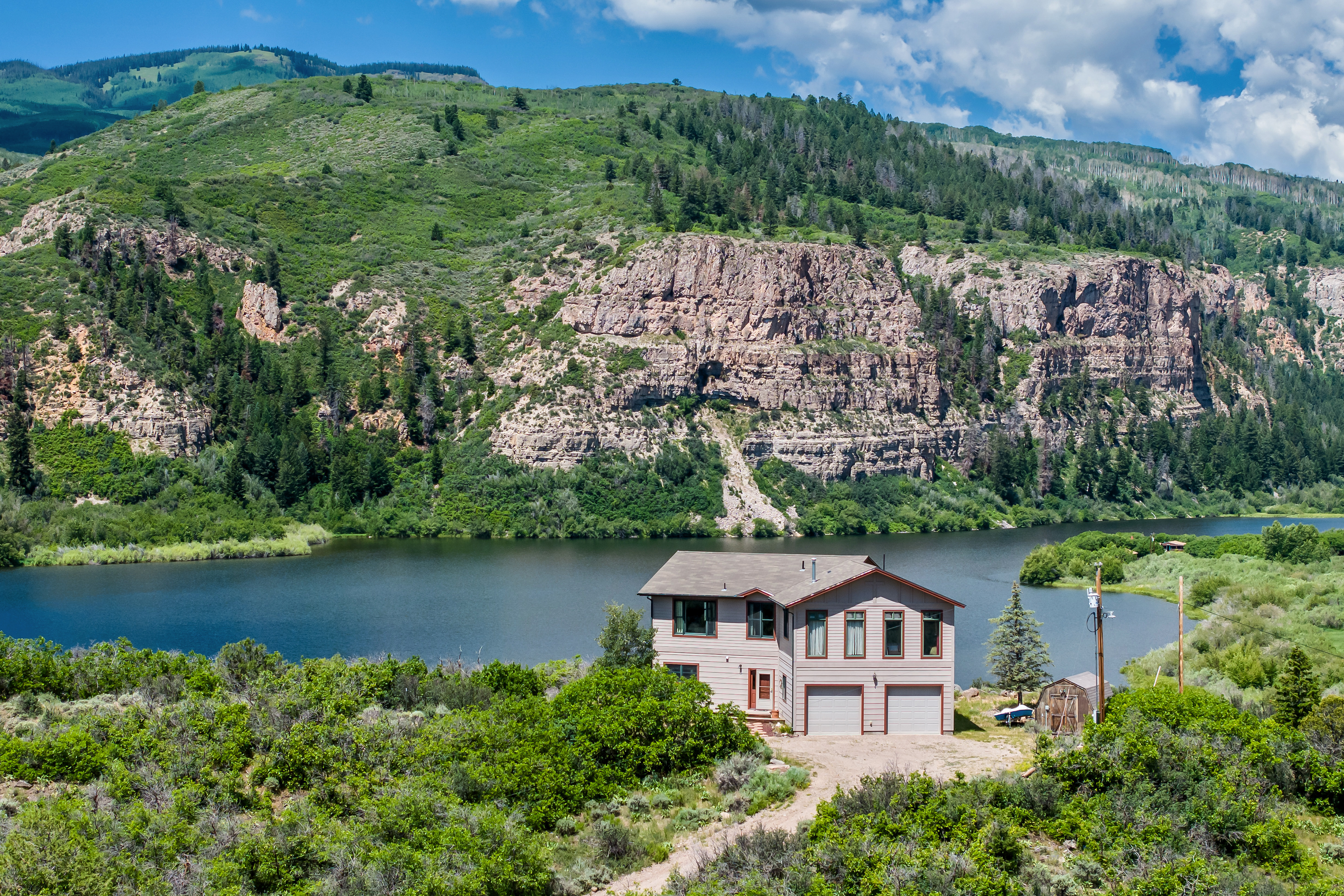 This aerial perspective captures a two-story residential home nestled in a serene, mountainous landscape beside a tranquil lake. The house features a neutral-toned exterior with a two-car garage, surrounded by lush green vegetation and dramatic rocky cliffs in the background. The composition emphasizes the property's secluded, scenic location, offering a peaceful and picturesque retreat.