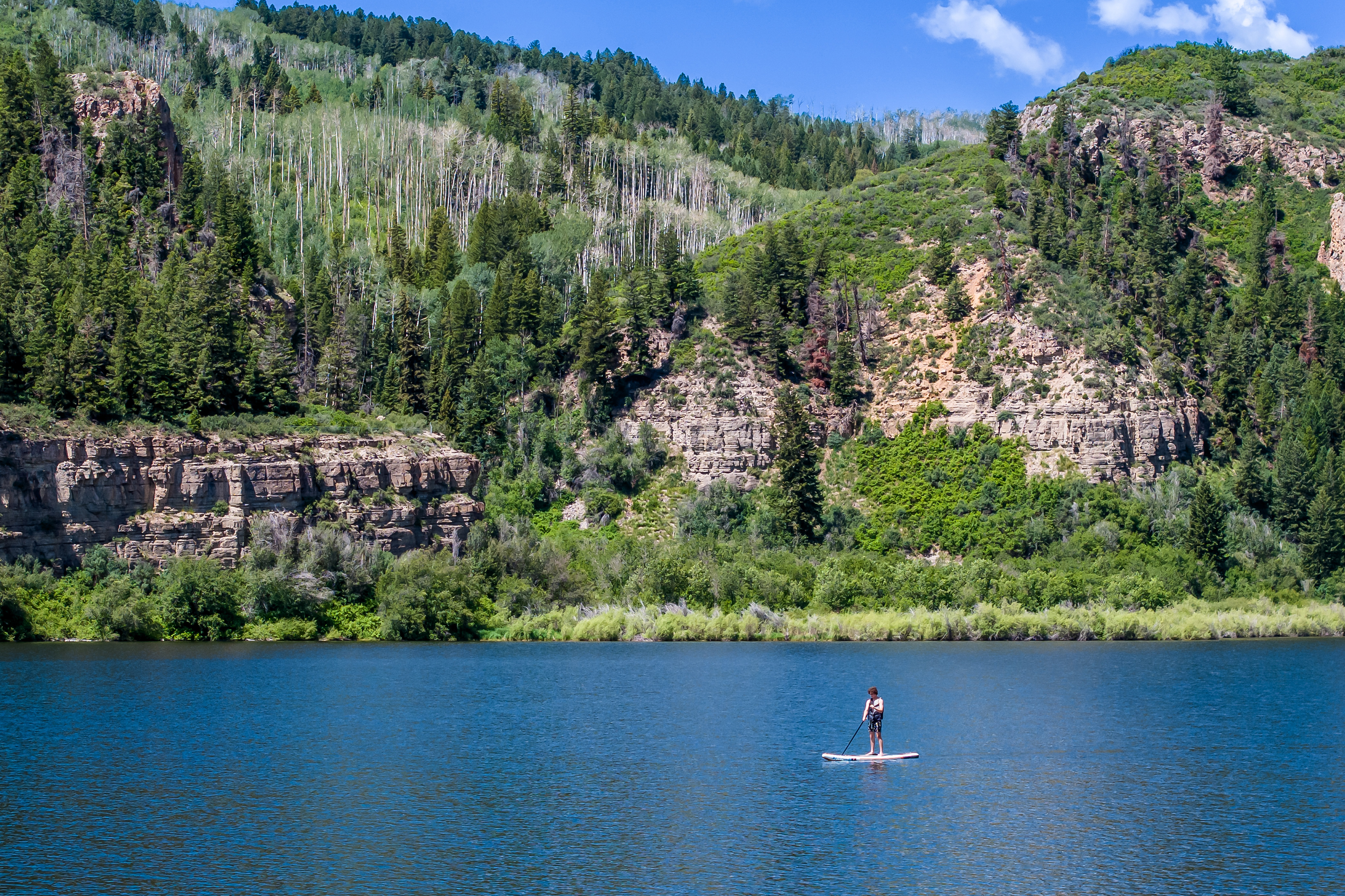 This scenic landscape shot captures a serene lake nestled at the base of a rugged, tree-covered mountain range. A lone individual is seen paddleboarding across the calm, deep blue water, emphasizing the recreational lifestyle opportunities available in the area. The composition highlights the natural beauty and tranquil environment, suggesting a peaceful retreat or outdoor-focused community setting.