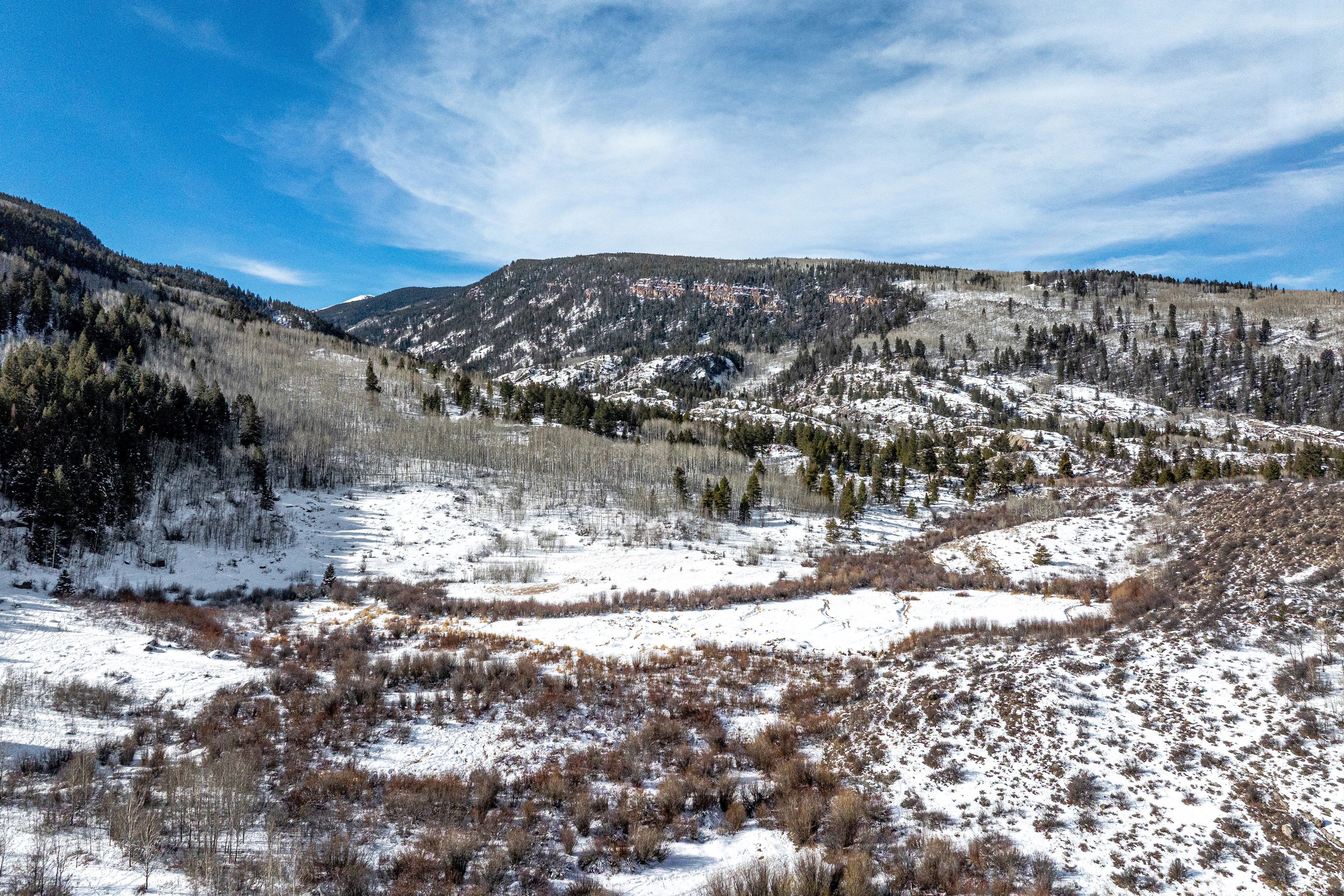 The image captures an aerial view of a snow-covered landscape, likely a mountain or valley area. There are trees dotted across the terrain, with a clear blue sky overhead. The image suggests a remote or secluded location, potentially suitable for a cabin or vacation home.