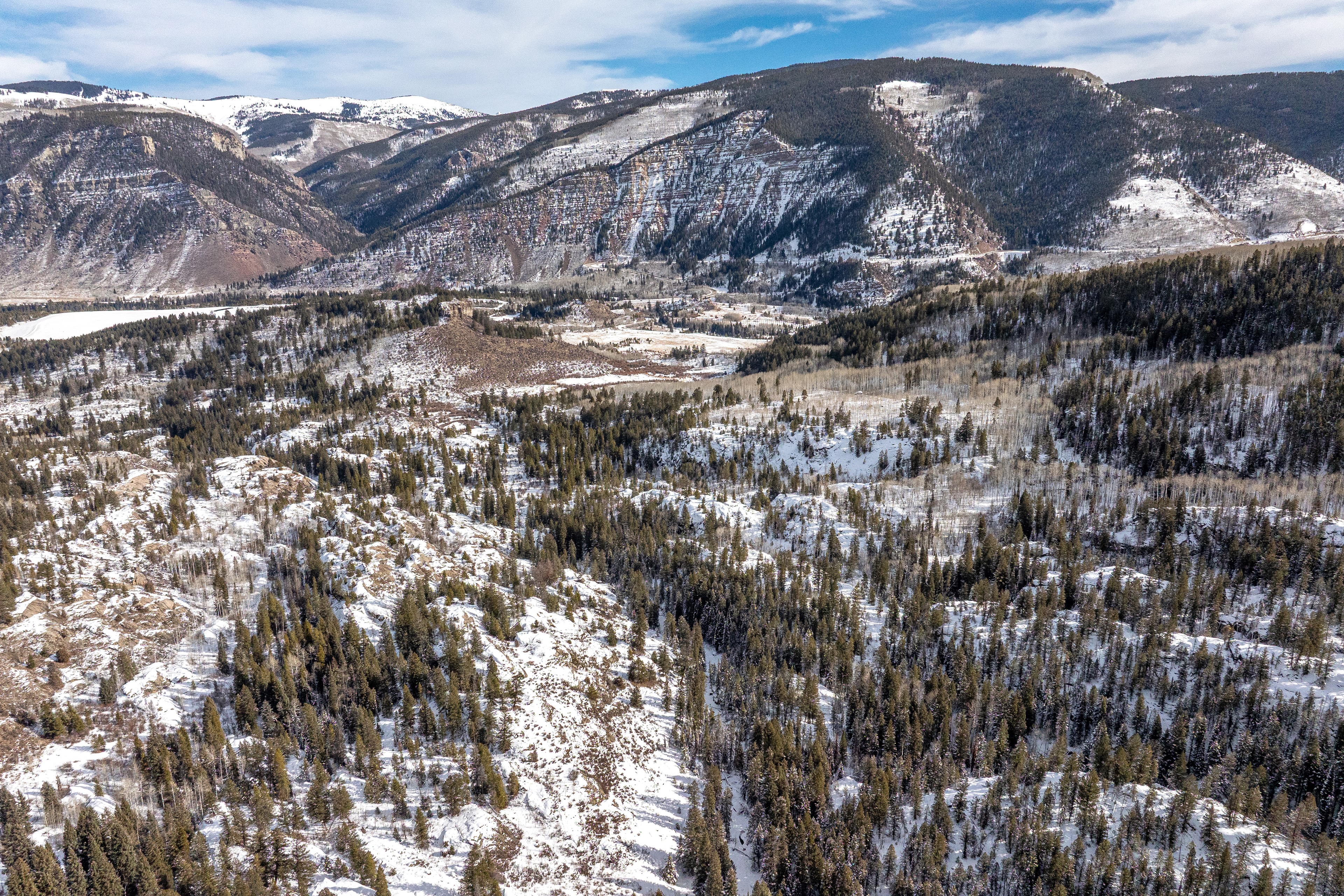 This aerial view showcases a mountainous landscape partially covered in snow. A dense forest fills the lower portions of the image, with a few open areas revealing snowy ground. The mountains in the background feature varied snow cover and rocky outcroppings, suggesting a remote and scenic location suitable for a secluded property.