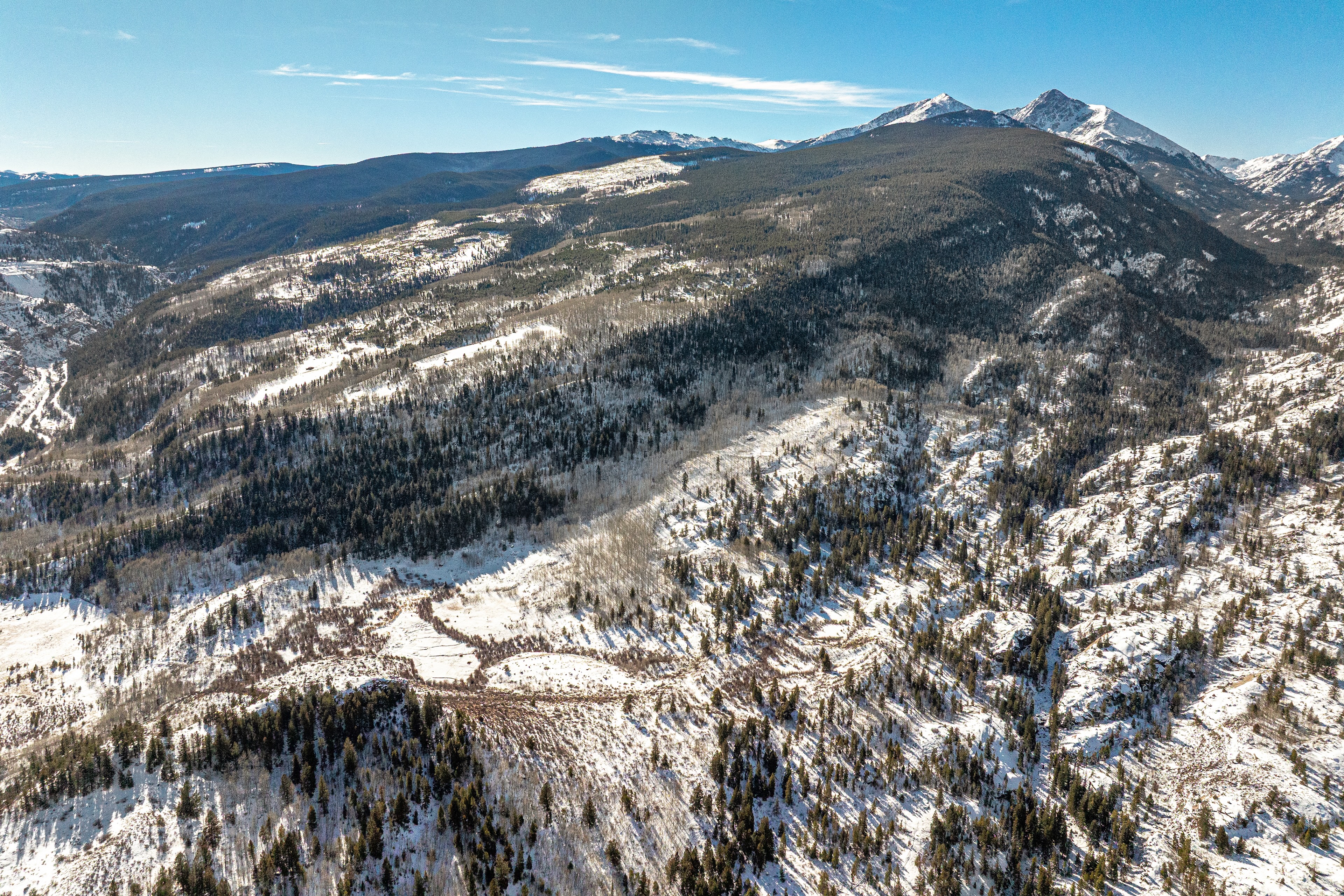 This is an aerial view of a mountain range in winter. The landscape is covered in snow, with trees visible on the valley floor and up the mountainsides. The sky is clear and blue, indicative of a beautiful day in this scenic, mountainous real estate location.