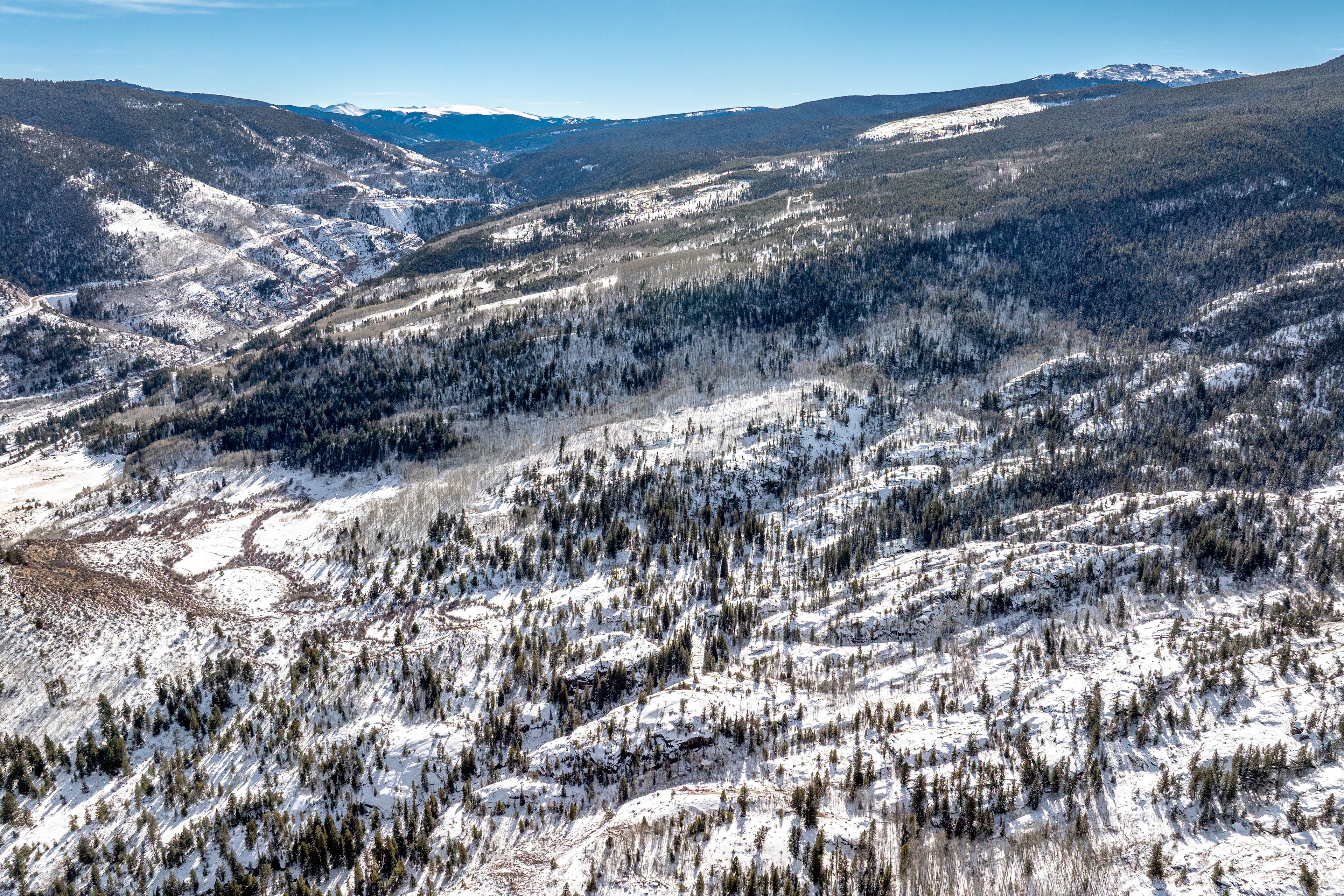 This aerial view captures a vast, mountainous landscape covered in snow and a mix of evergreen and deciduous trees. The image showcases the natural beauty and potential privacy of a secluded property. The clear blue sky contrasts with the snowy terrain, creating a scenic and appealing vista.