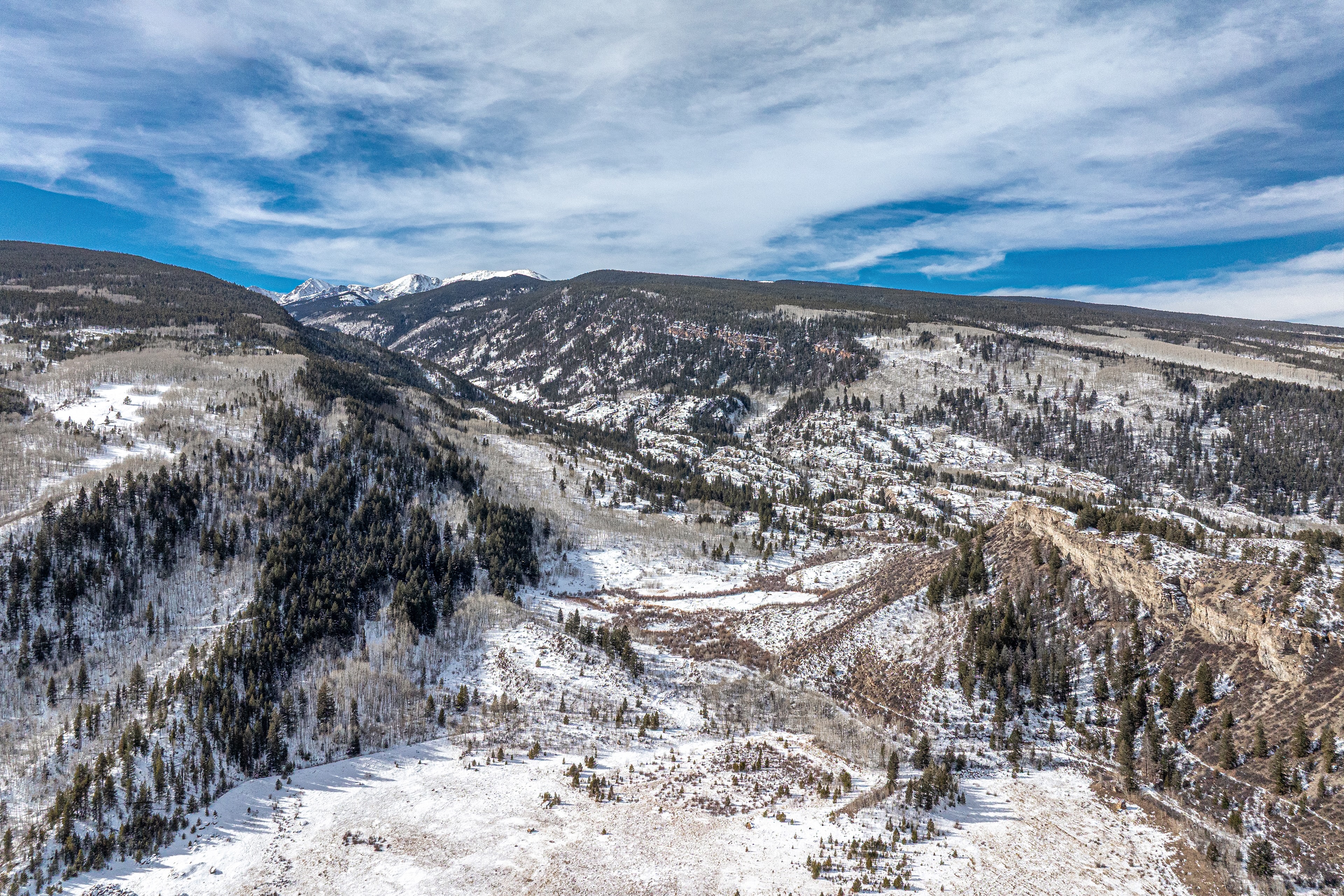This is an aerial view of a mountainous landscape in winter. The mountains are partially covered in snow and a mix of evergreen and bare deciduous trees. The sky is blue with scattered clouds, creating a scenic and natural environment that may appeal to buyers seeking a tranquil and remote property.