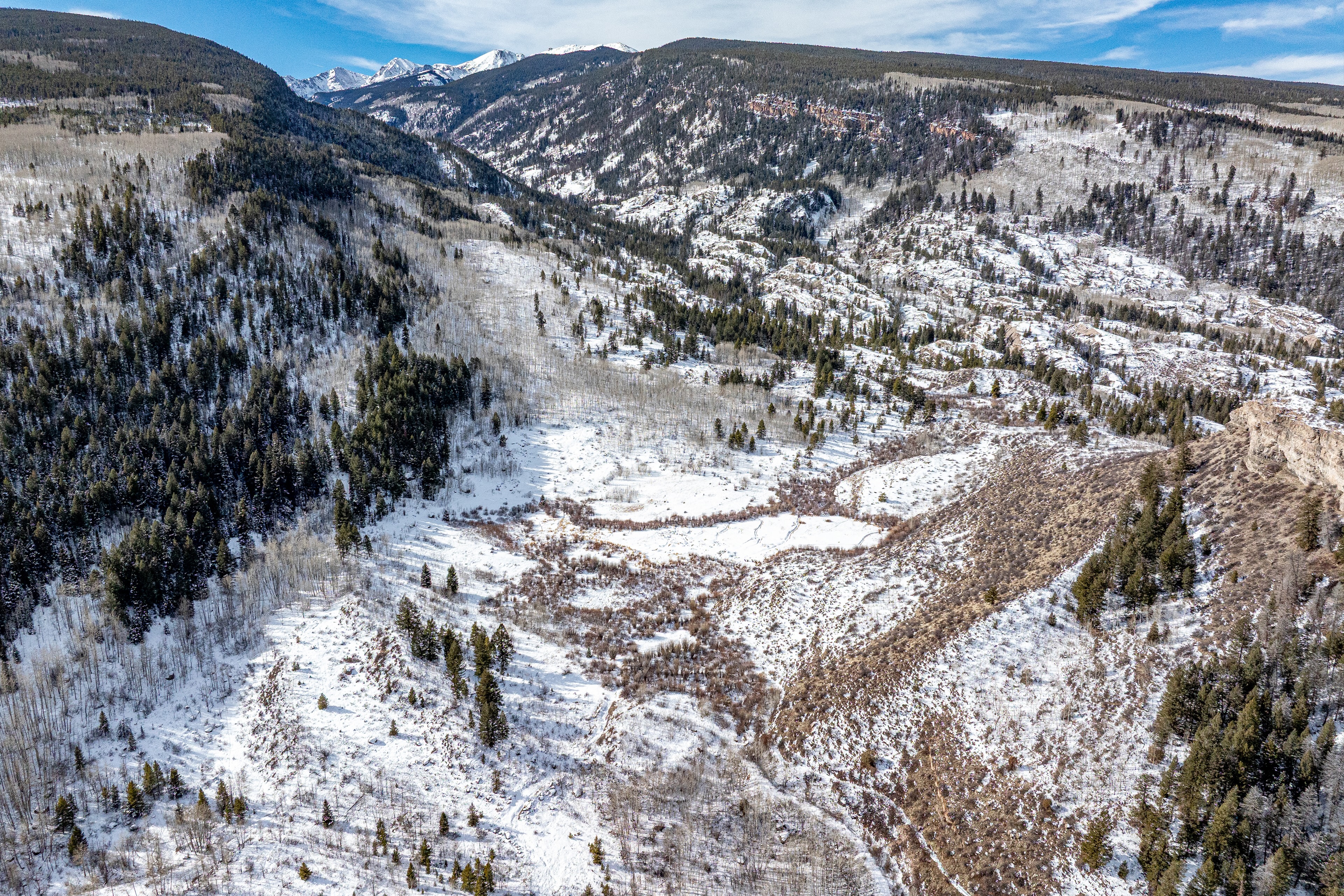 This is an aerial view of a mountainous landscape, likely taken in winter. Snow covers the ground and trees, creating a picturesque scene. The terrain appears rugged and natural, potentially appealing to buyers interested in secluded properties or scenic views.