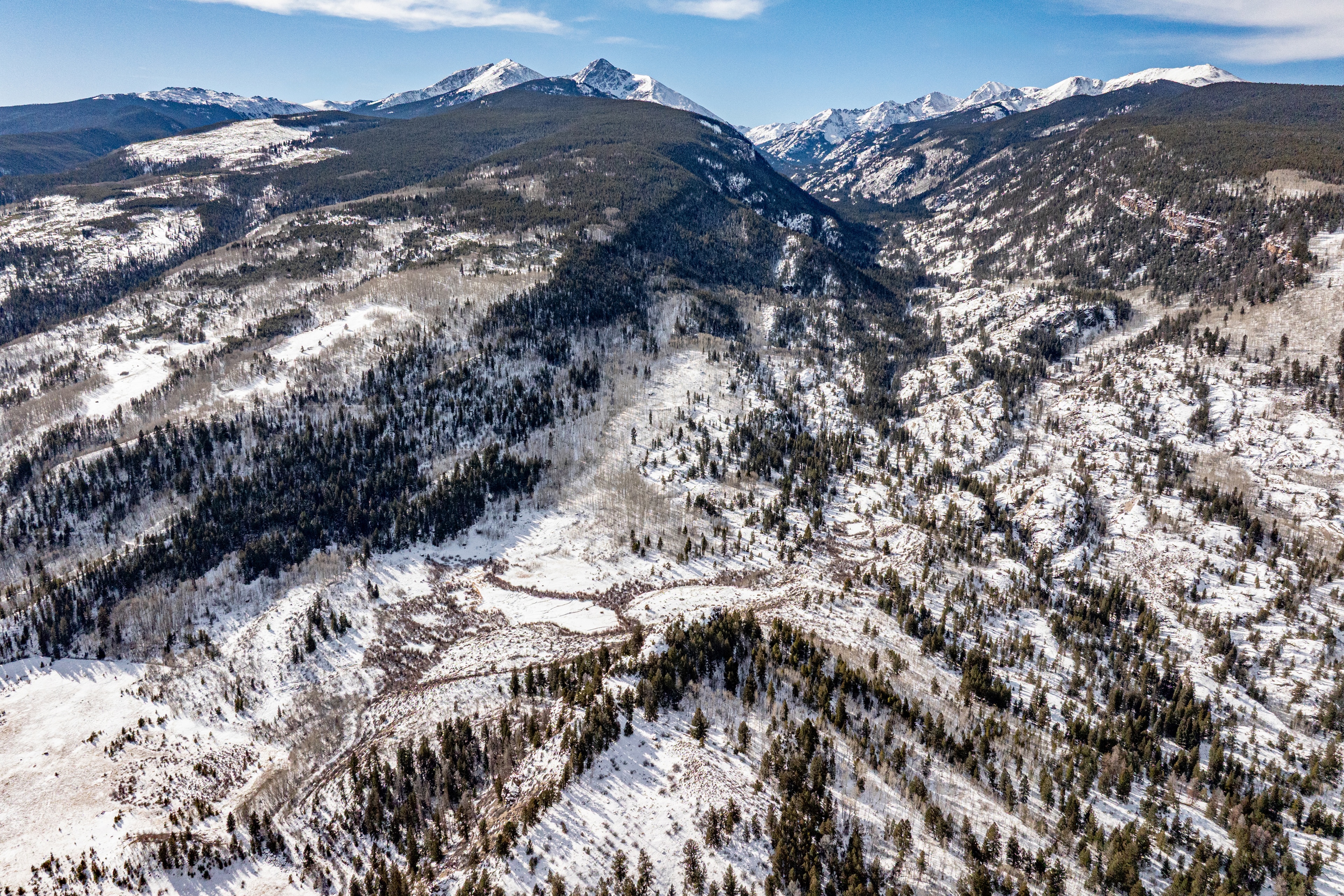 This aerial view showcases a vast mountain landscape covered in patches of snow and dense forests. The rugged terrain adds a sense of seclusion and natural beauty, potentially highlighting the property's privacy and proximity to outdoor recreational opportunities. The clear sky suggests a desirable location for enjoying the natural environment.