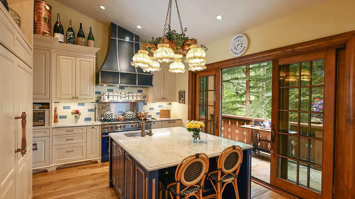 This is a well-appointed traditional kitchen with cream-colored cabinetry, a contrasting blue island with a marble countertop, and stainless steel appliances. A decorative chandelier hangs above the island. The kitchen also features an adjacent outdoor patio area accessible through large glass doors, bringing natural light and a connection to the outdoors.