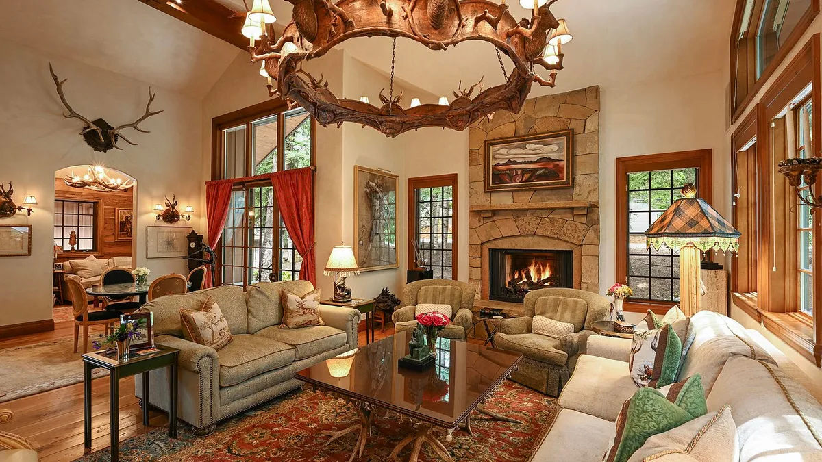 This is a luxurious living room featuring a large stone fireplace, comfortable seating, and a unique antler chandelier. The room is decorated in a classic, rustic style with warm colors and natural materials. Large windows provide ample natural light and views of the surrounding landscape.
