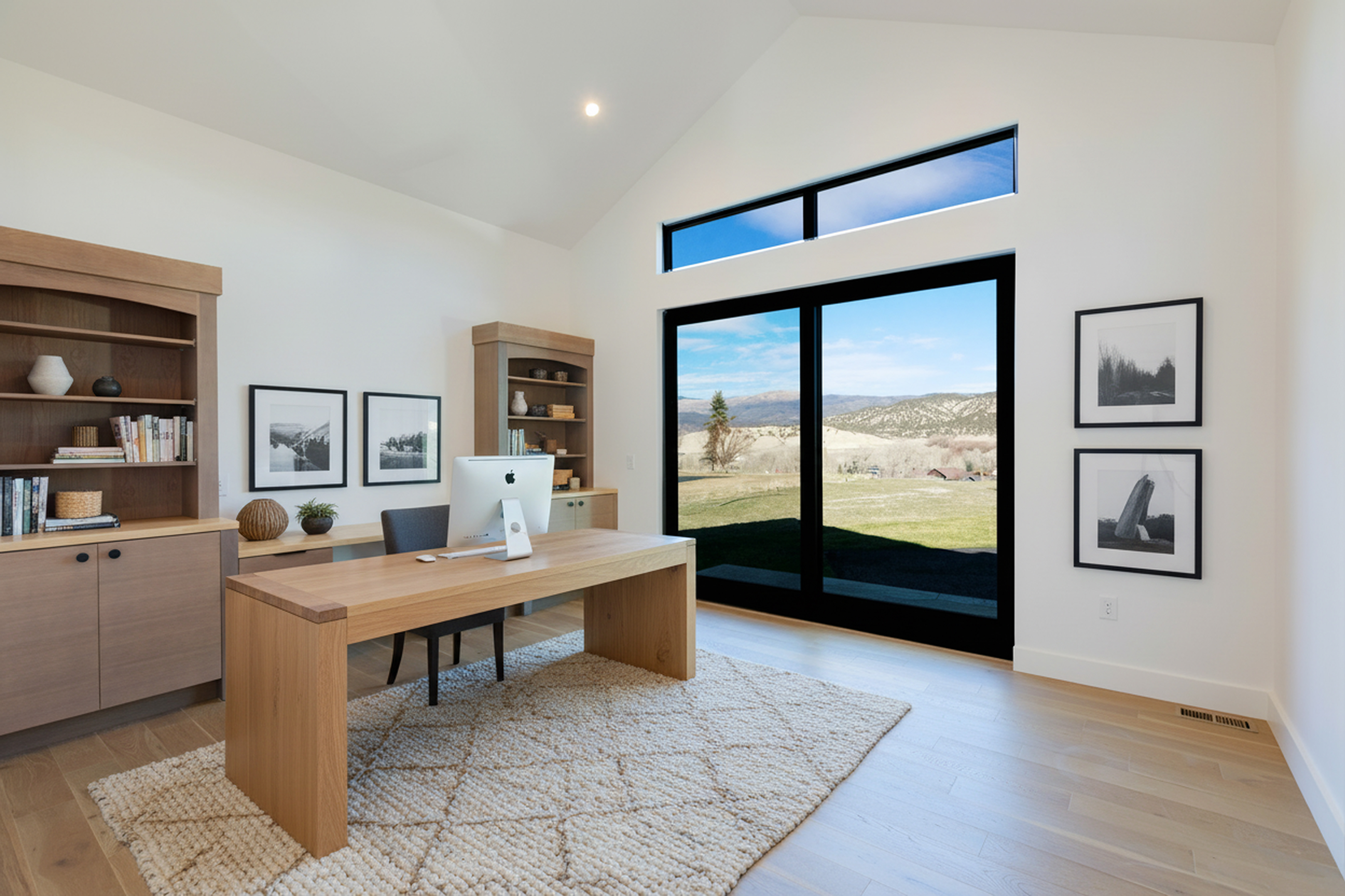 This bright and airy home office features built-in shelving and a large desk. Natural light floods the space through a large window, offering scenic views. The room is well-organized and designed for productivity, creating an inviting workspace.