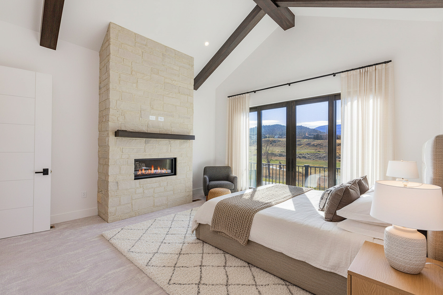 This is the primary bedroom of a home. It features a stone fireplace, white walls, and dark wood architectural beams. A large window/door offers a view of the landscape. The bed is neatly made, and there is a nightstand with a lamp.