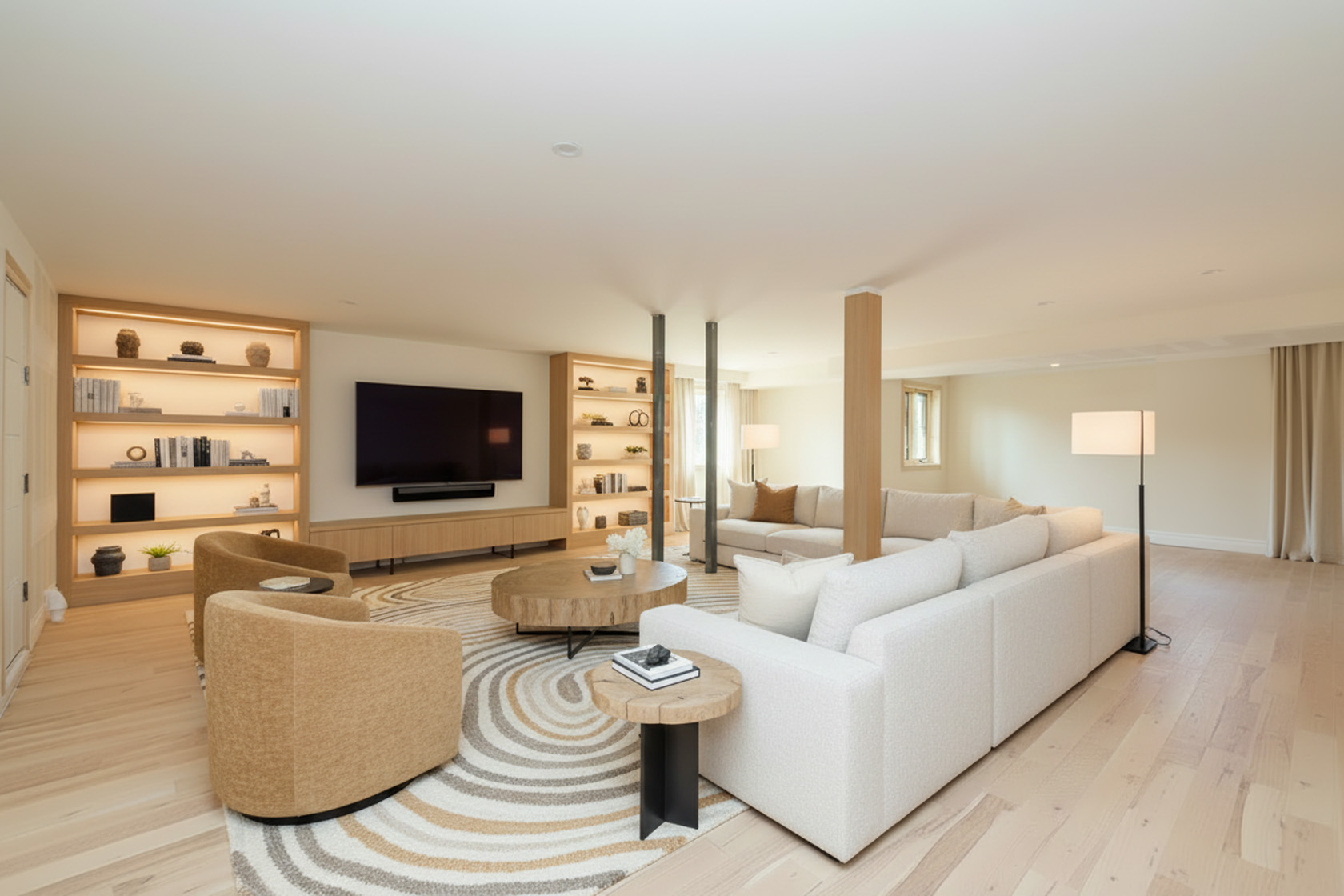 This inviting living room features a neutral color palette with warm wood accents. The space is anchored by a patterned circular rug and showcases a comfortable seating arrangement with multiple sofas and armchairs. Built-in shelving with accent lighting adds a touch of sophistication and provides ample storage.