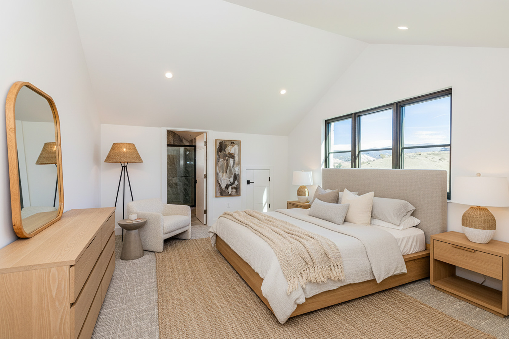 This is a bright and airy primary bedroom with vaulted ceilings and a neutral color palette. The room features a large bed with a padded headboard, light wood nightstands, and a coordinating dresser with a full-length mirror. A sitting area with an armchair and a floor lamp adds to the room's comfort and functionality. The abundant natural light from the window highlights the serene atmosphere, making it an attractive space for potential buyers.