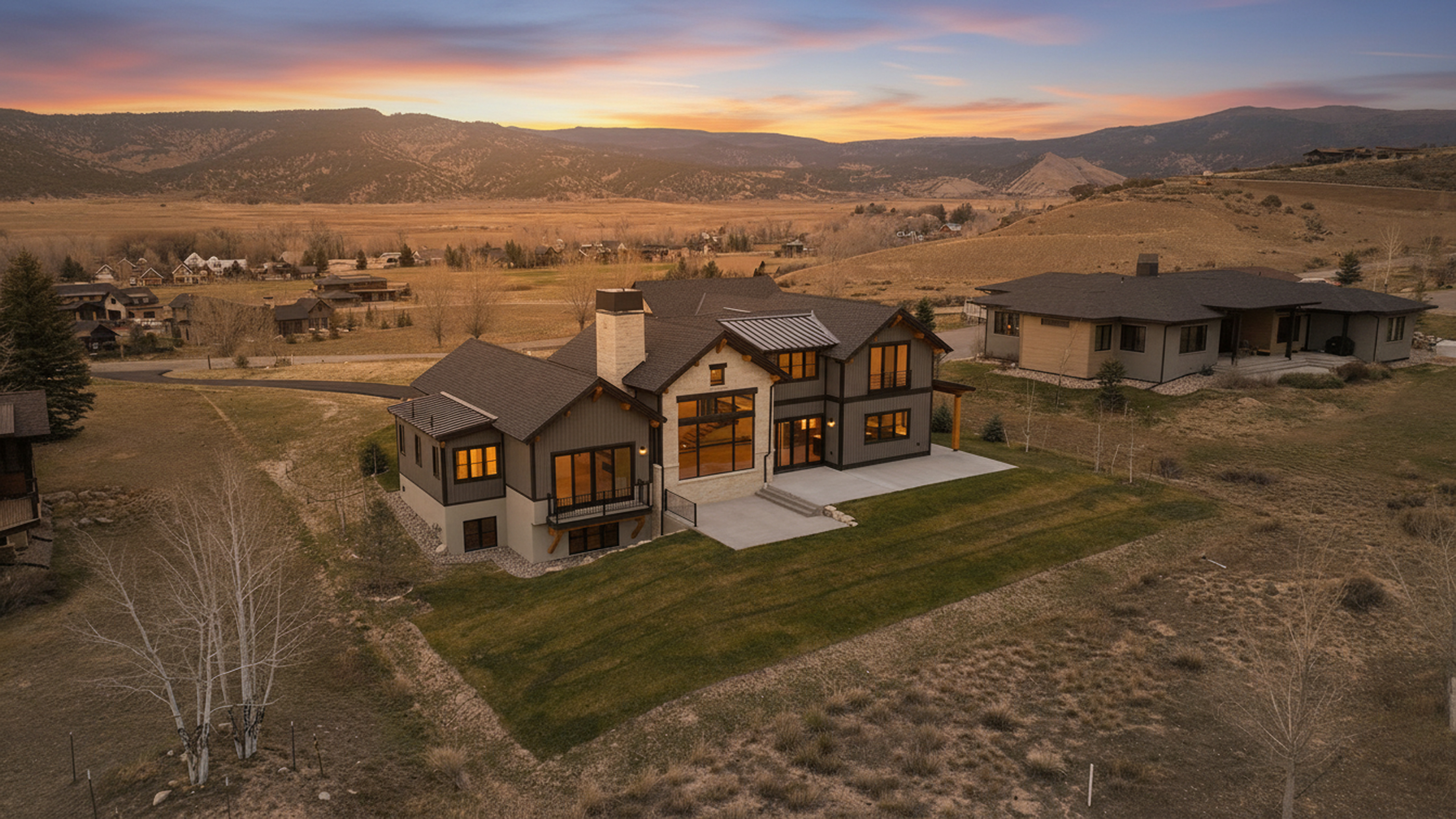 This aerial shot features a modern two-story home with dark gray siding, light stone accents, and a dark roof. A well-maintained lawn surrounds the house, contrasting with the natural dry landscape. The residence is situated in a spacious lot, with other homes visible in the distance, all set against a scenic backdrop of mountains and a striking sunset sky.
