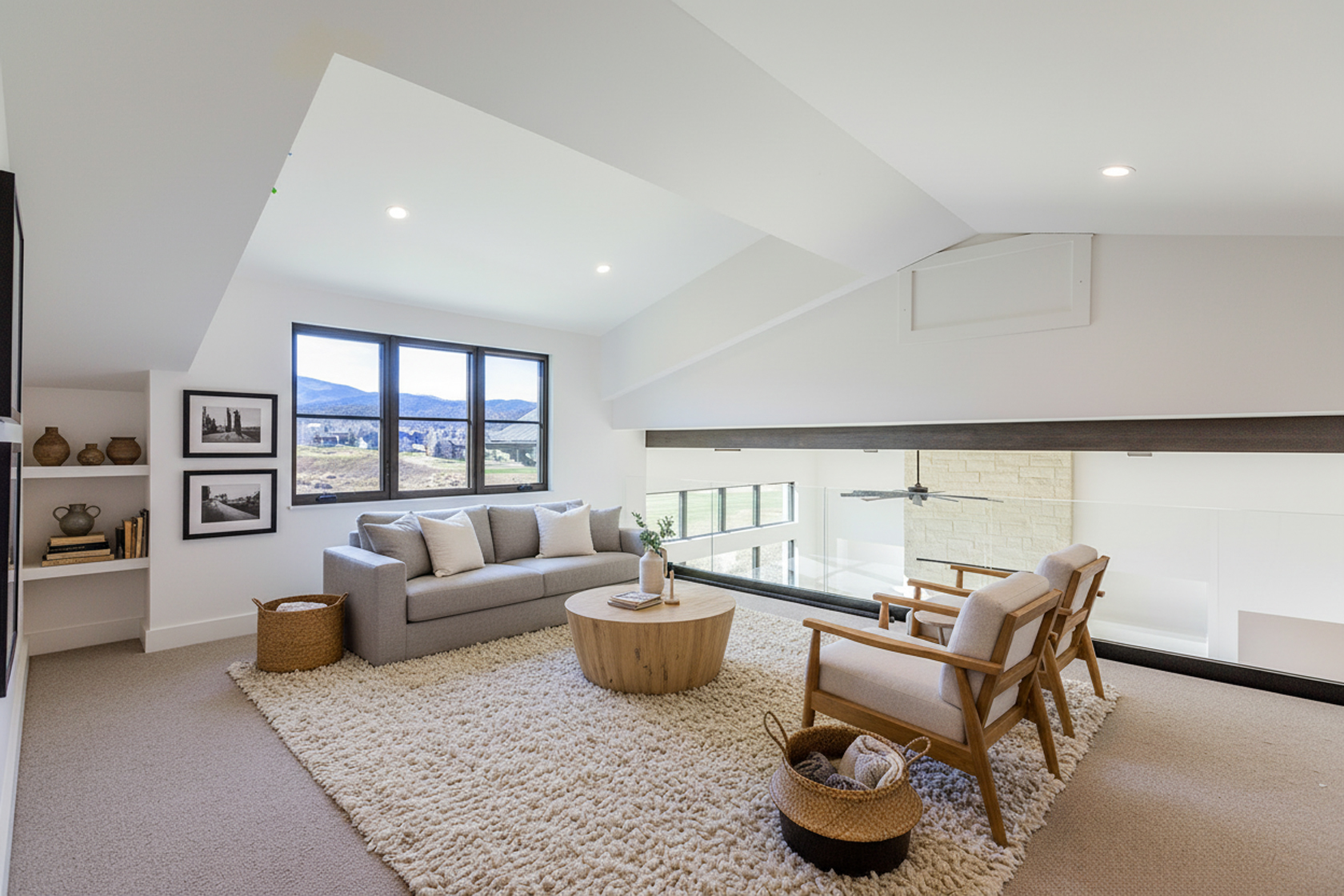 This is a bright and airy living room featuring a modern design. The space includes a comfortable gray sofa, a round wooden coffee table, and two armchairs, all situated on a plush rug. Large windows provide ample natural light and views of the surrounding landscape.