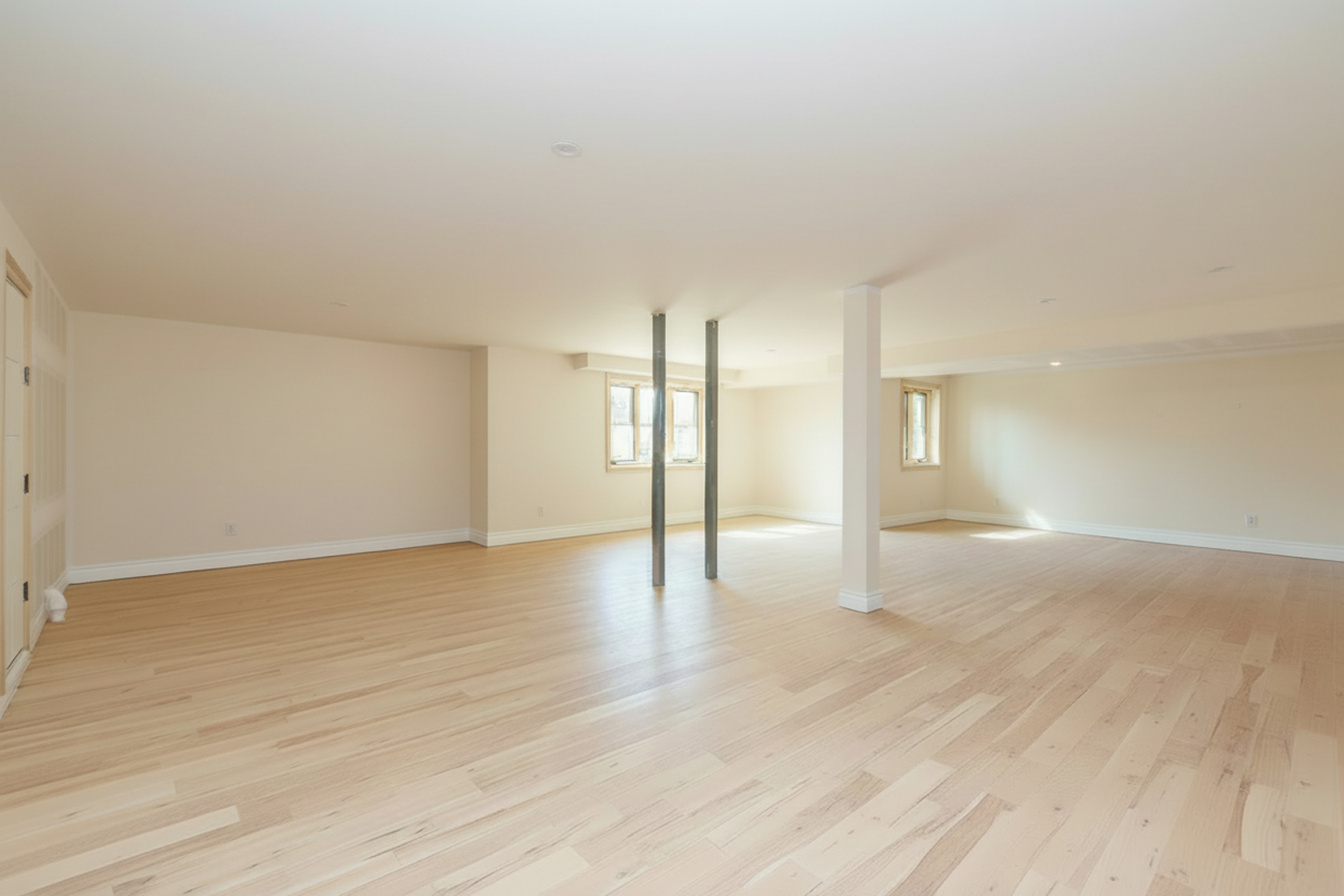 This is a spacious, unfinished basement with light-colored wooden flooring and neutral walls. Natural light streams through the windows, highlighting the open floor plan. Support beams are visible, suggesting potential for customization and finishing to suit various needs.