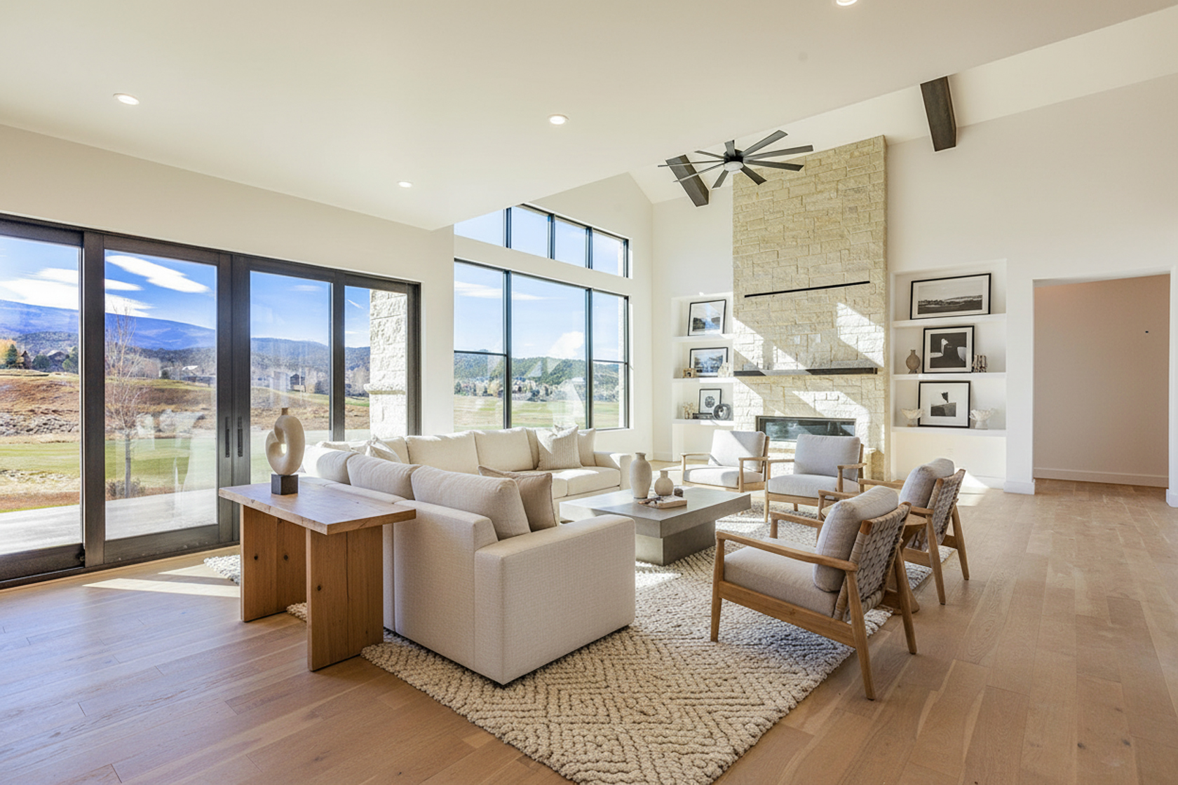 This spacious living room features a neutral color palette, hardwood floors, and a textured rug. Large windows provide ample natural light and beautiful outdoor views. A stone fireplace adds a focal point, and modern furniture creates a comfortable and stylish atmosphere.
