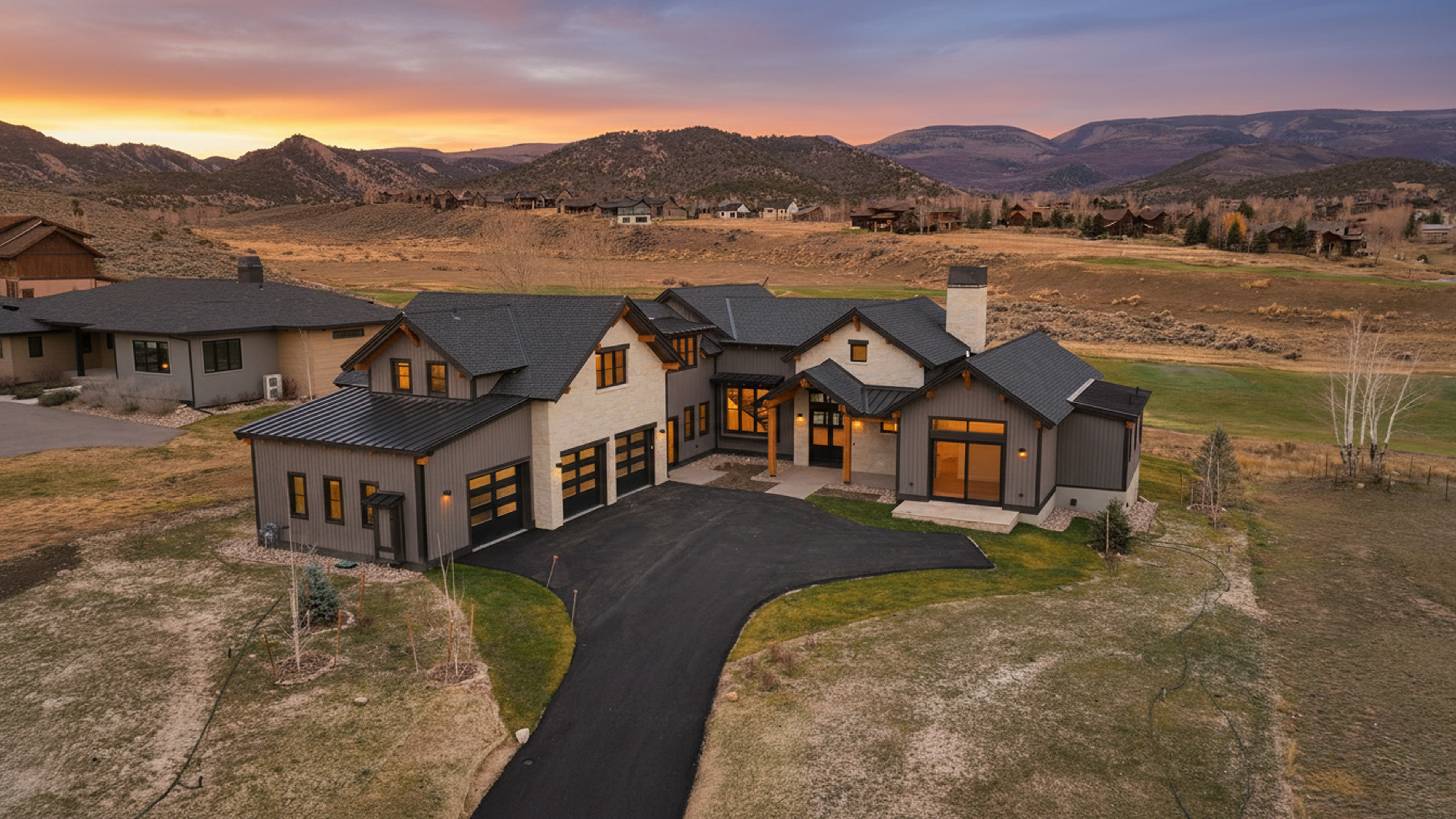 This aerial view showcases a beautifully designed home with a dark-colored roof and a combination of light and dark exterior finishes. The property features an expansive yard and a paved driveway leading to a two-car garage. The house is situated in a community with scenic mountain views, adding to its appeal.