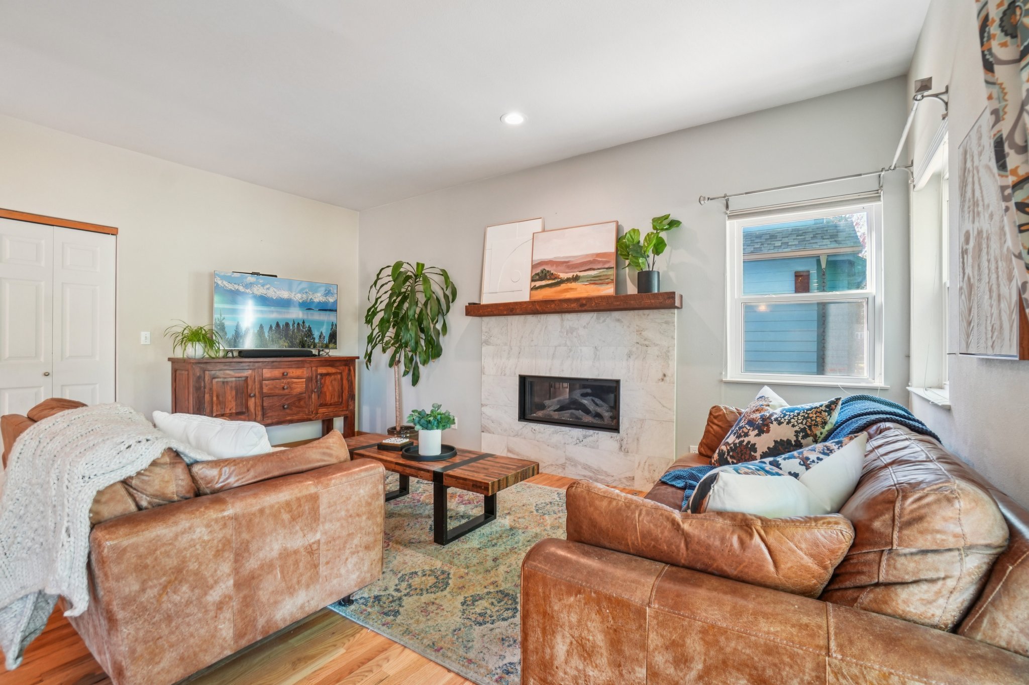 This is a cozy living room featuring leather sofas, a marble fireplace with a wooden mantel, and a wooden console table with a TV. The room is decorated with plants and artwork, creating a warm and inviting atmosphere. The perspective is from the center of the room, capturing the fireplace and seating area.