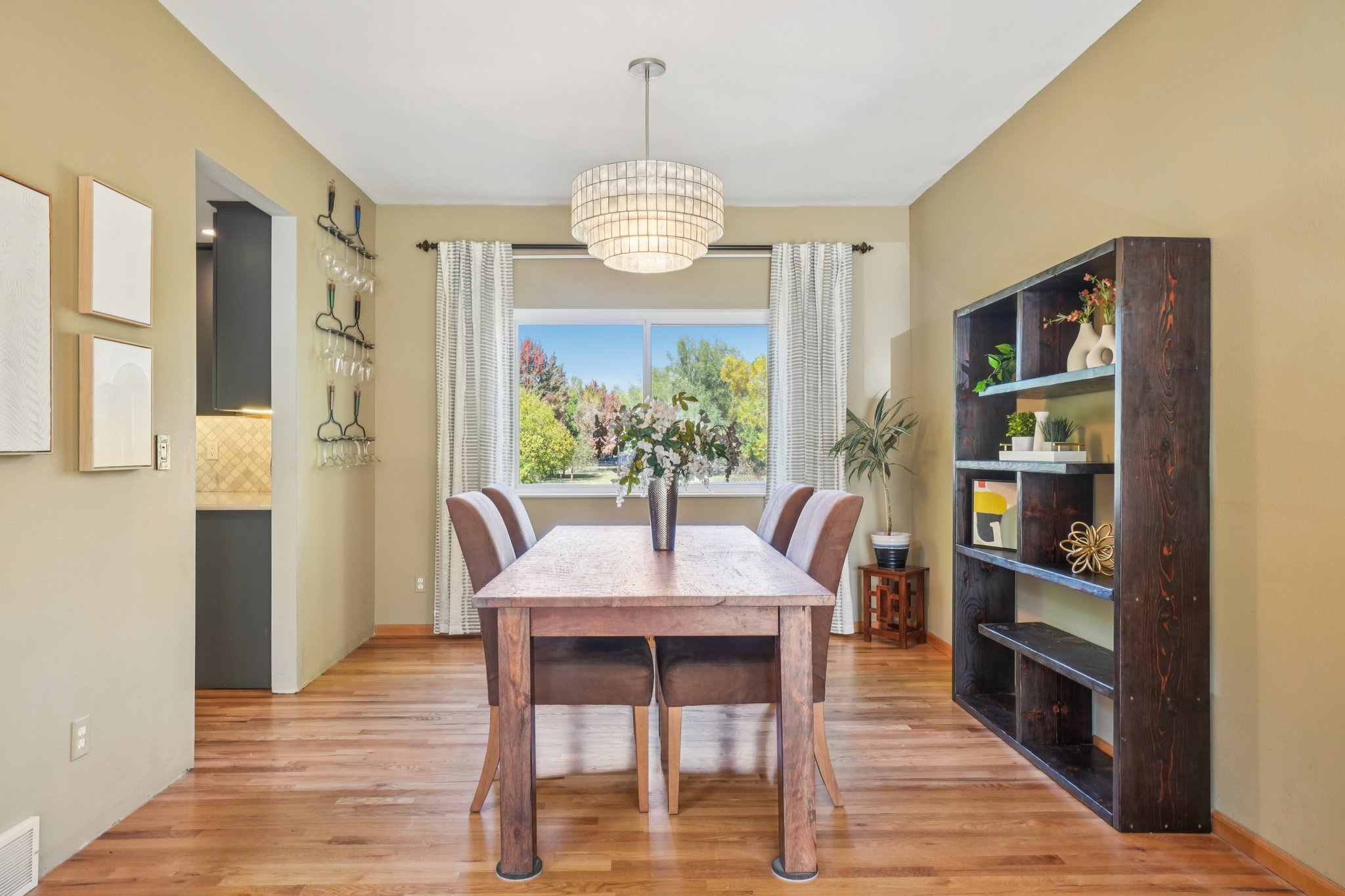 This is an interior shot of a dining room featuring a wooden table with chairs, a modern chandelier, and a large window providing natural light. A dark wood bookshelf stands against the wall, adding character to the space. The room has a warm and inviting atmosphere, perfect for family meals and gatherings.
