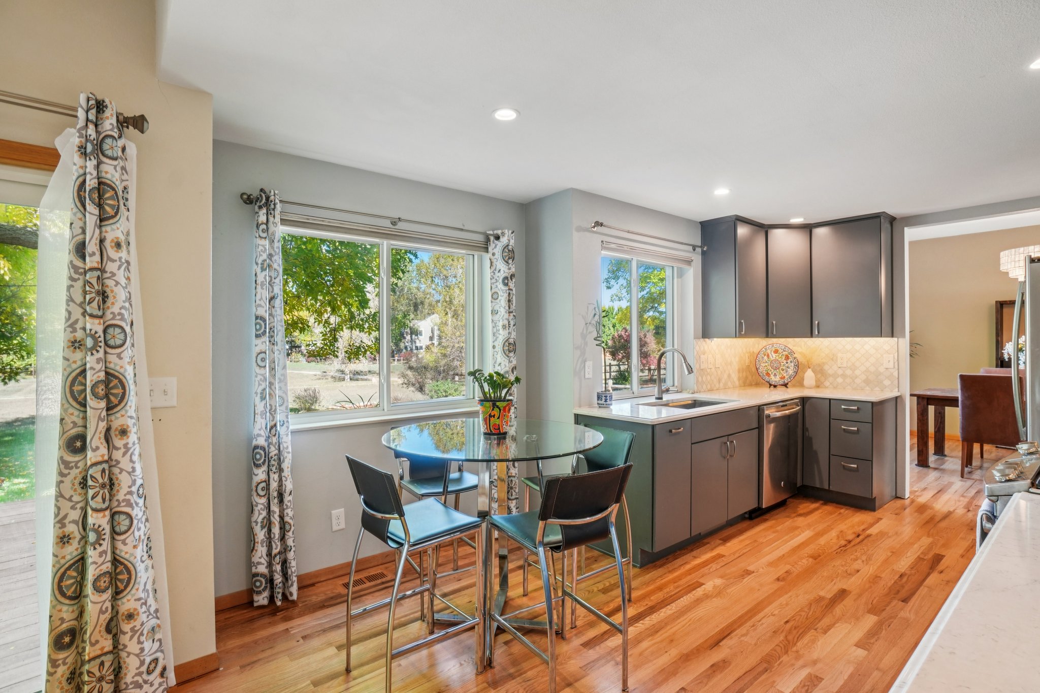 This is a bright and modern kitchen featuring gray cabinetry, stainless steel appliances, and hardwood flooring. A glass-top table with bar stools sits near a window, providing a casual dining area. The kitchen is well-lit with recessed lighting and natural light, creating a welcoming and functional space.