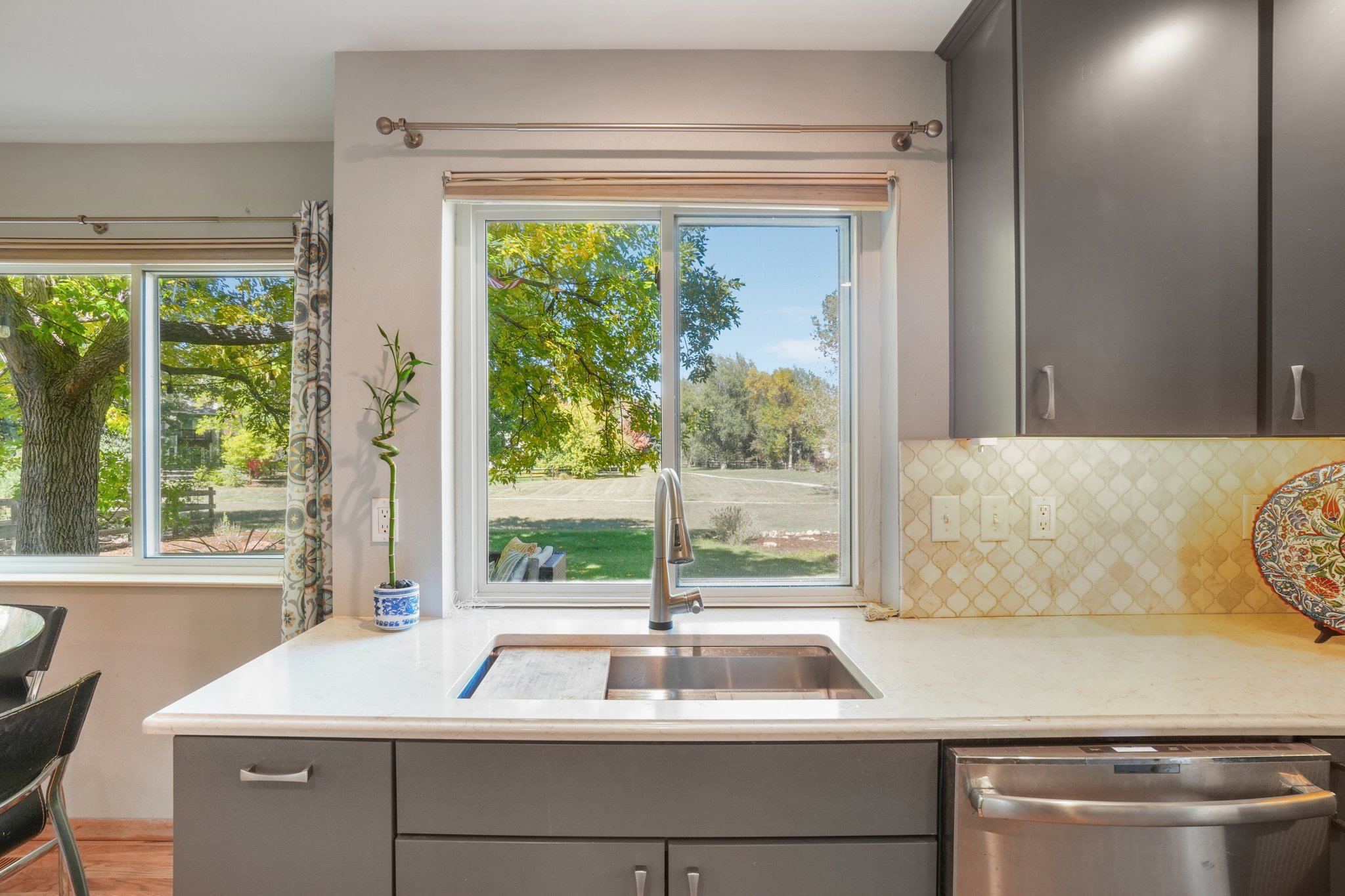 This is a well-lit kitchen featuring gray cabinetry, stainless steel appliances, and a white countertop with a built-in sink. A large window above the sink offers a view of the outdoors, bringing natural light into the space. The backsplash is a light-colored mosaic tile, adding a touch of elegance.