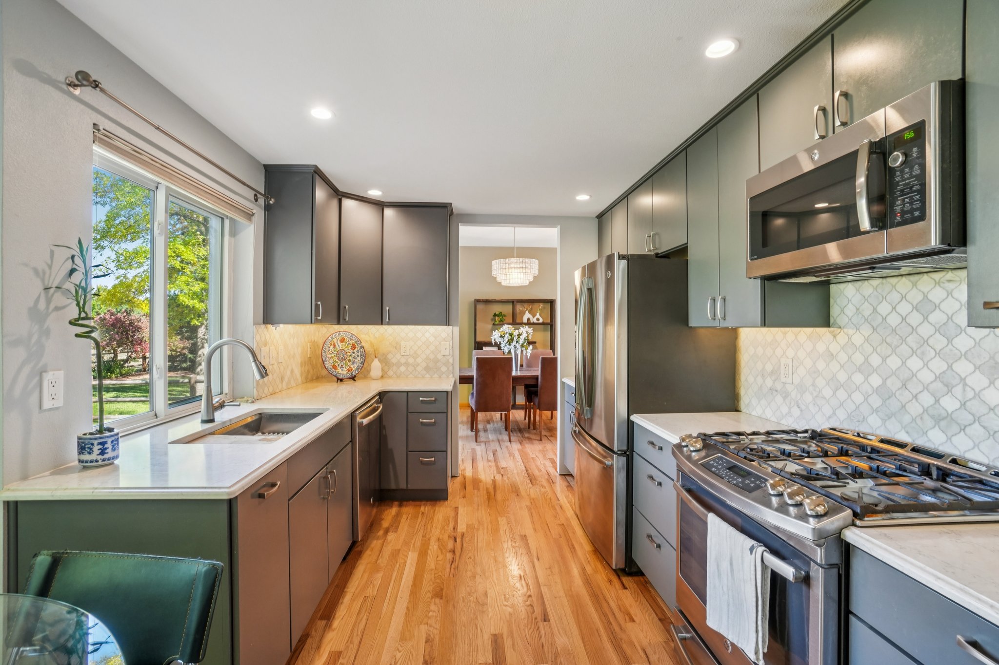 This is a well-lit kitchen featuring dark gray cabinetry, stainless steel appliances, and light-colored countertops. The hardwood flooring adds warmth to the space, and a window provides natural light. The kitchen has a modern and functional design, with a view into the dining area.