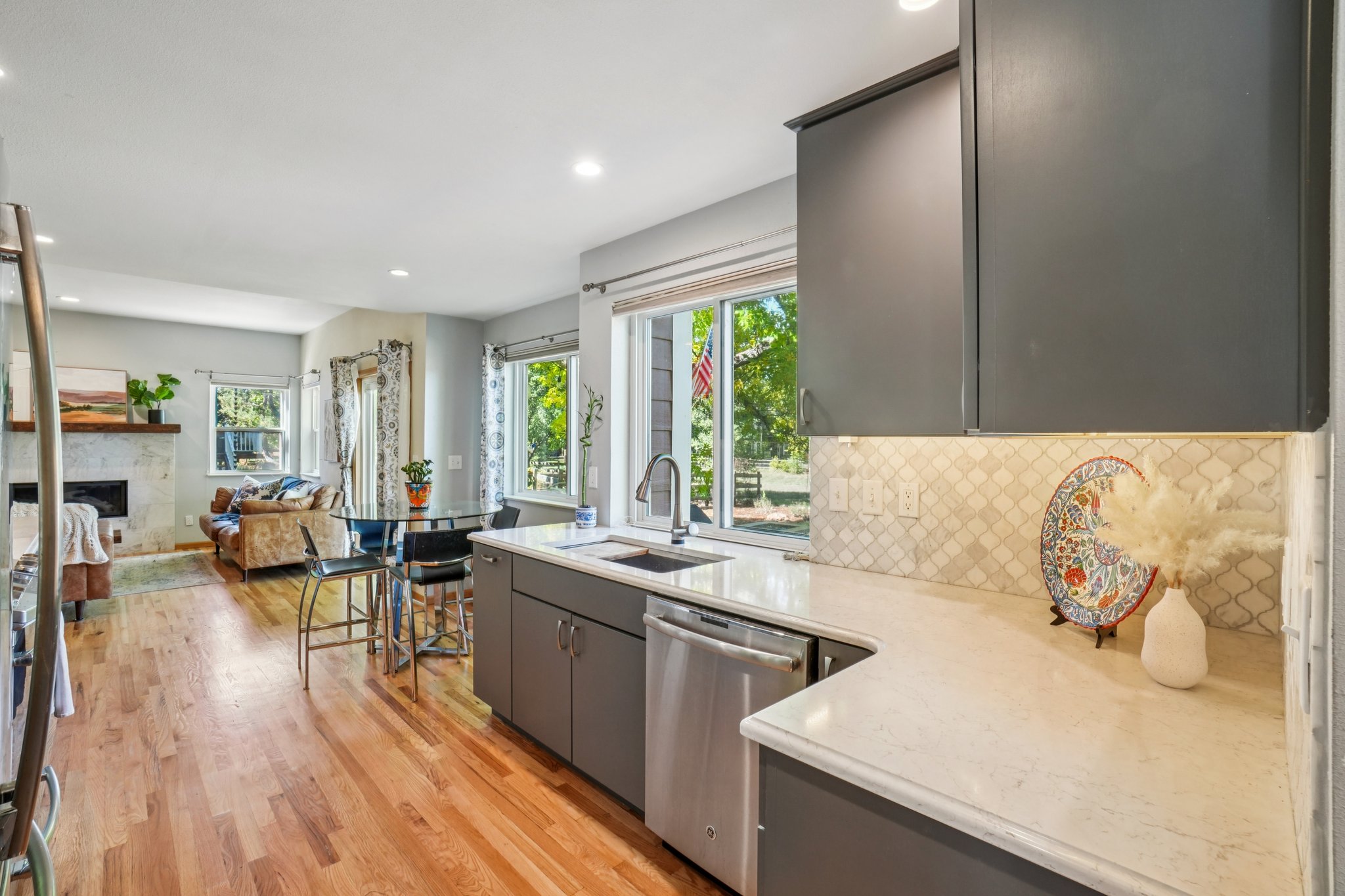 This is a bright and airy kitchen with hardwood floors and gray cabinetry. The countertops are a light quartz, and the backsplash is a decorative tile. Stainless steel appliances and a large window over the sink add to the modern feel of the space, which opens to a living area.