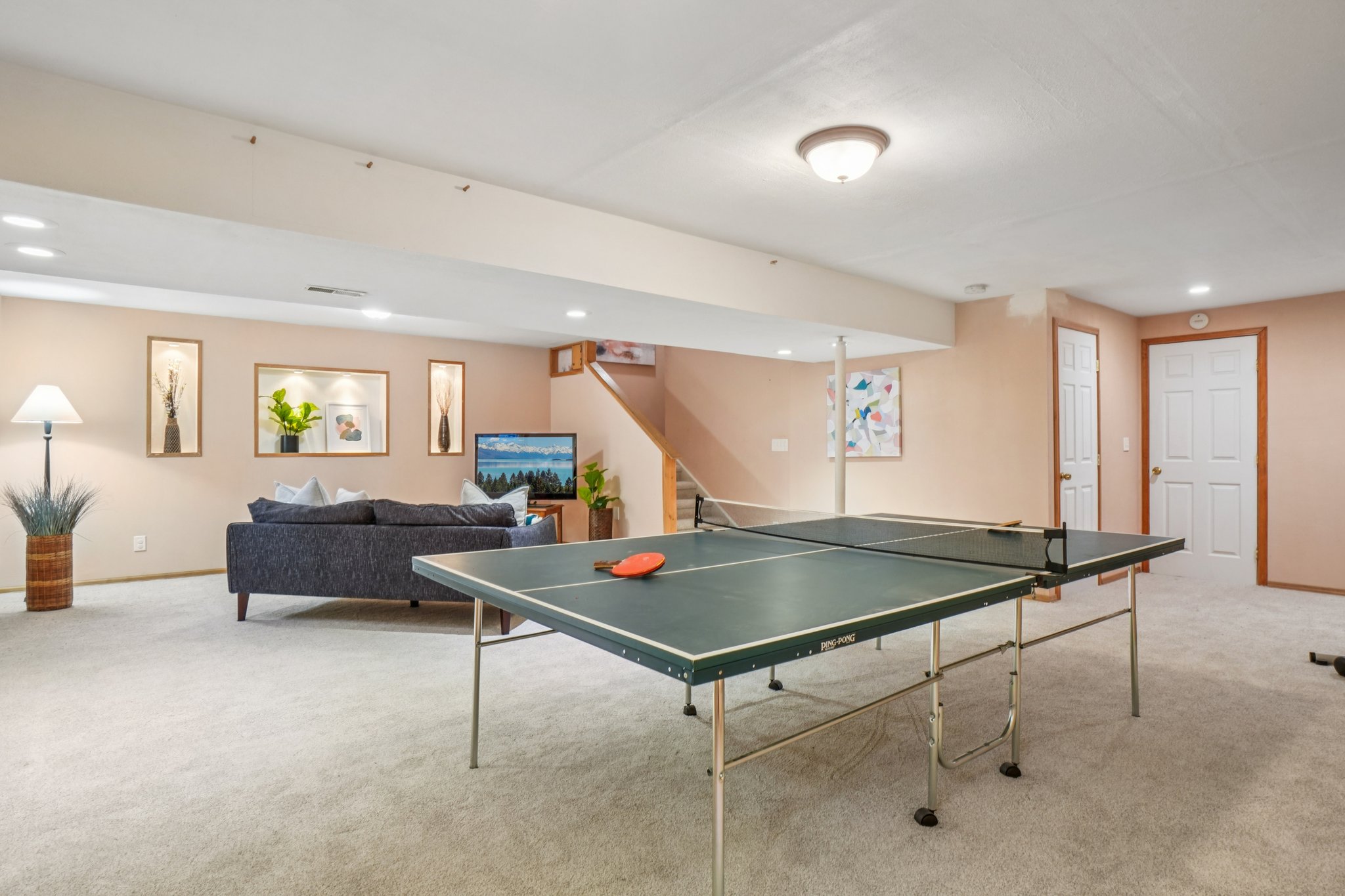 This is a spacious basement featuring a ping pong table in the foreground and a comfortable living area in the background. The living area includes a gray sofa, framed artwork, and a television. The basement is well-lit with recessed lighting and has neutral-toned walls and carpet, creating a versatile and inviting space.