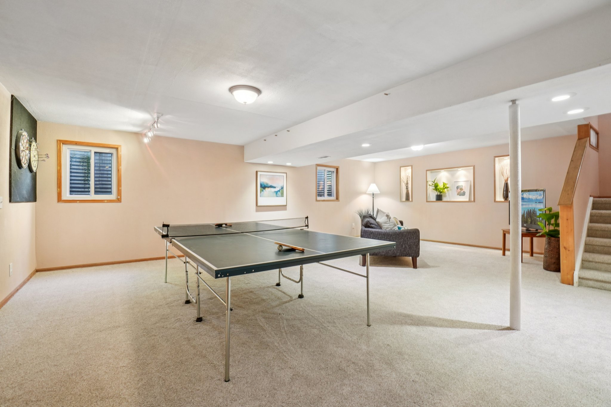 This is a spacious finished basement featuring a ping pong table as a focal point, suggesting a recreational area. The room is carpeted and has neutral-colored walls, with recessed lighting and windows providing illumination. A seating area with a sofa and a staircase leading to the upper level are also visible, indicating a comfortable and functional living space.