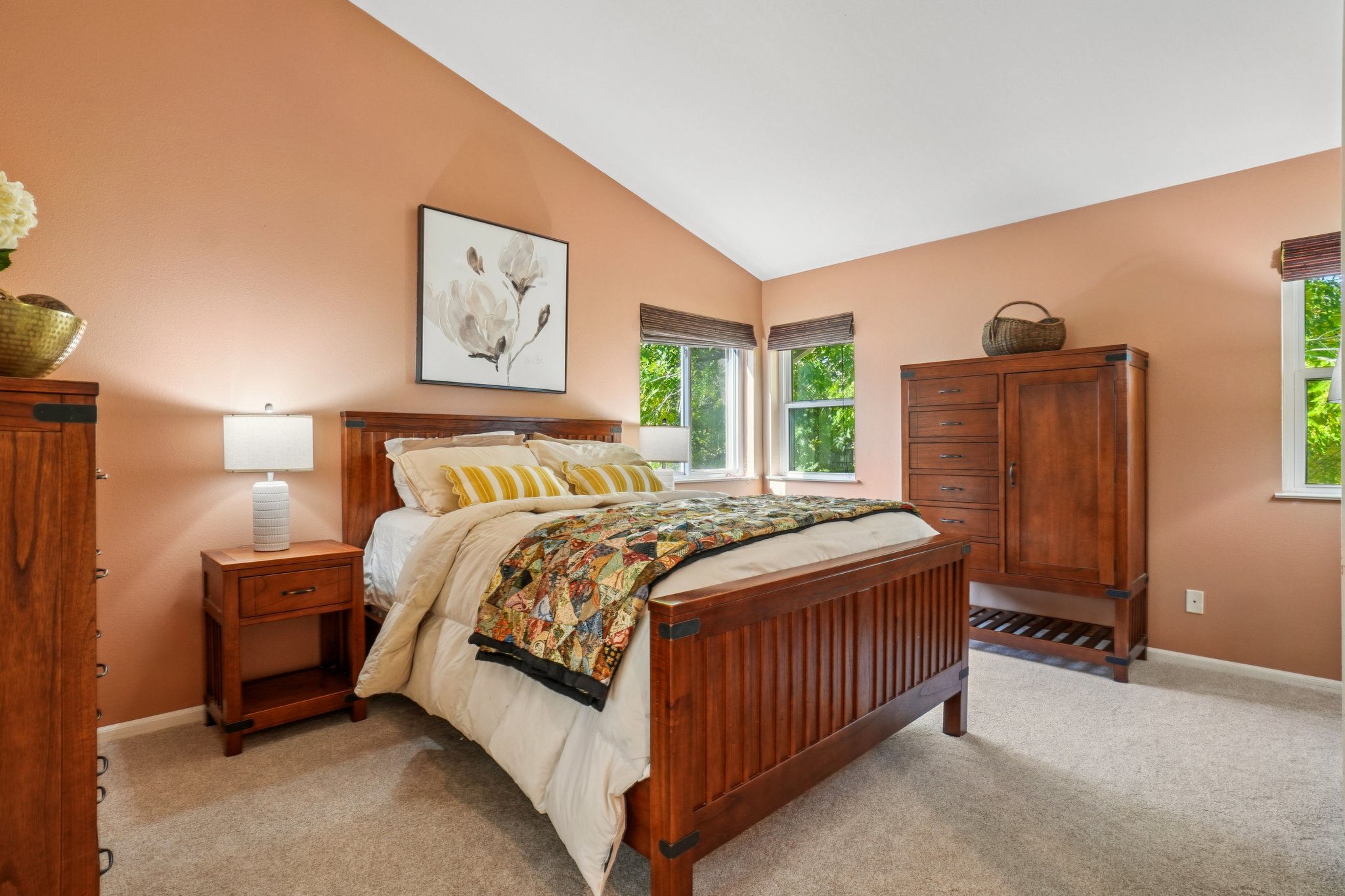 This is a cozy primary bedroom featuring a wooden bed frame with a patterned quilt and yellow striped pillows. The room has a warm, inviting feel with peach-colored walls, natural light from the windows, and wooden furniture including a nightstand with a lamp and a tall dresser. A framed floral print hangs above the bed, adding a touch of elegance.