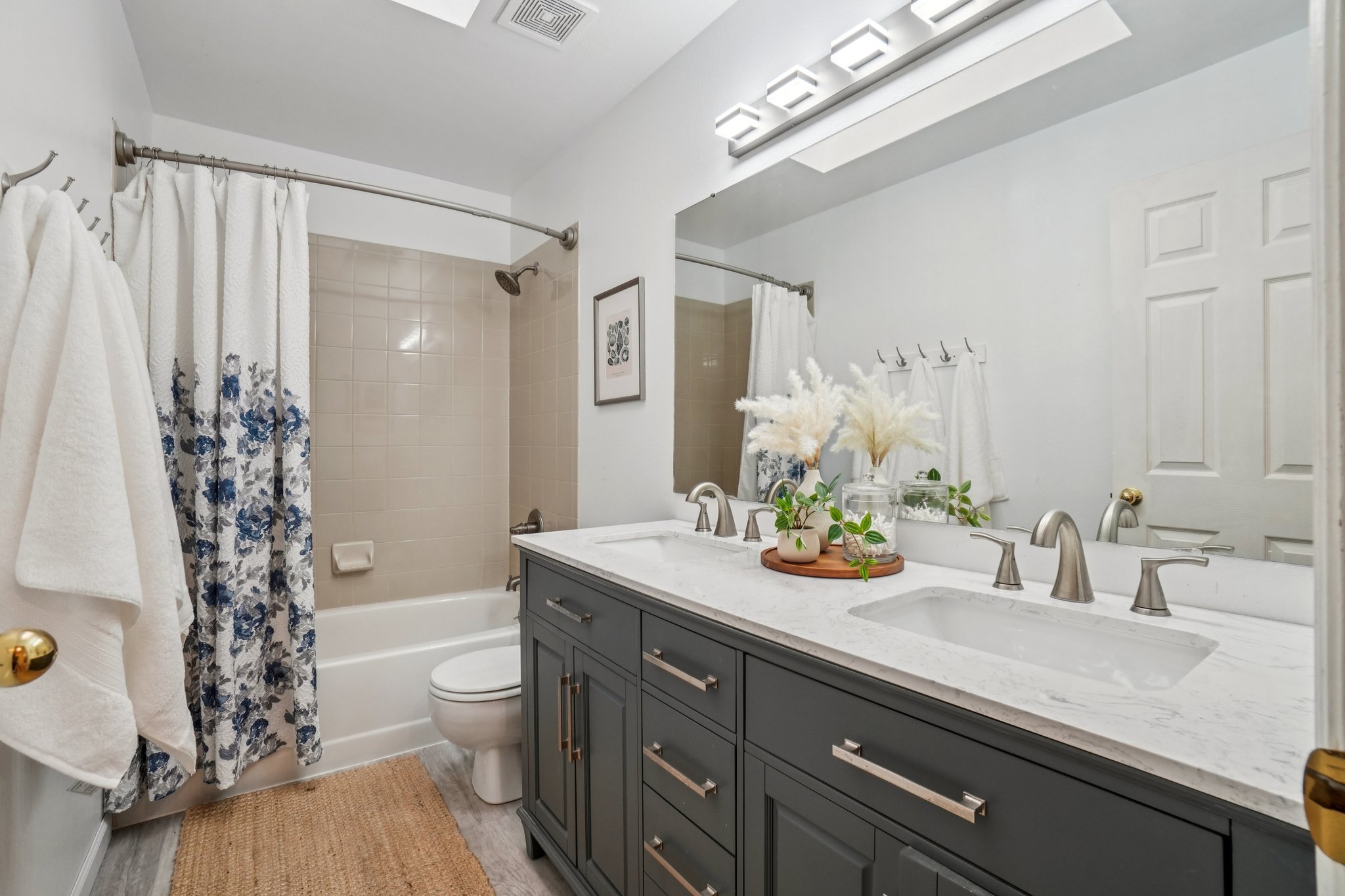 This is a well-lit bathroom featuring a double vanity with a gray cabinet and white countertop. The bathroom includes a shower-tub combination with a floral shower curtain, a toilet, and a neutral-toned rug. The overall impression is clean and functional, with a touch of decorative flair.