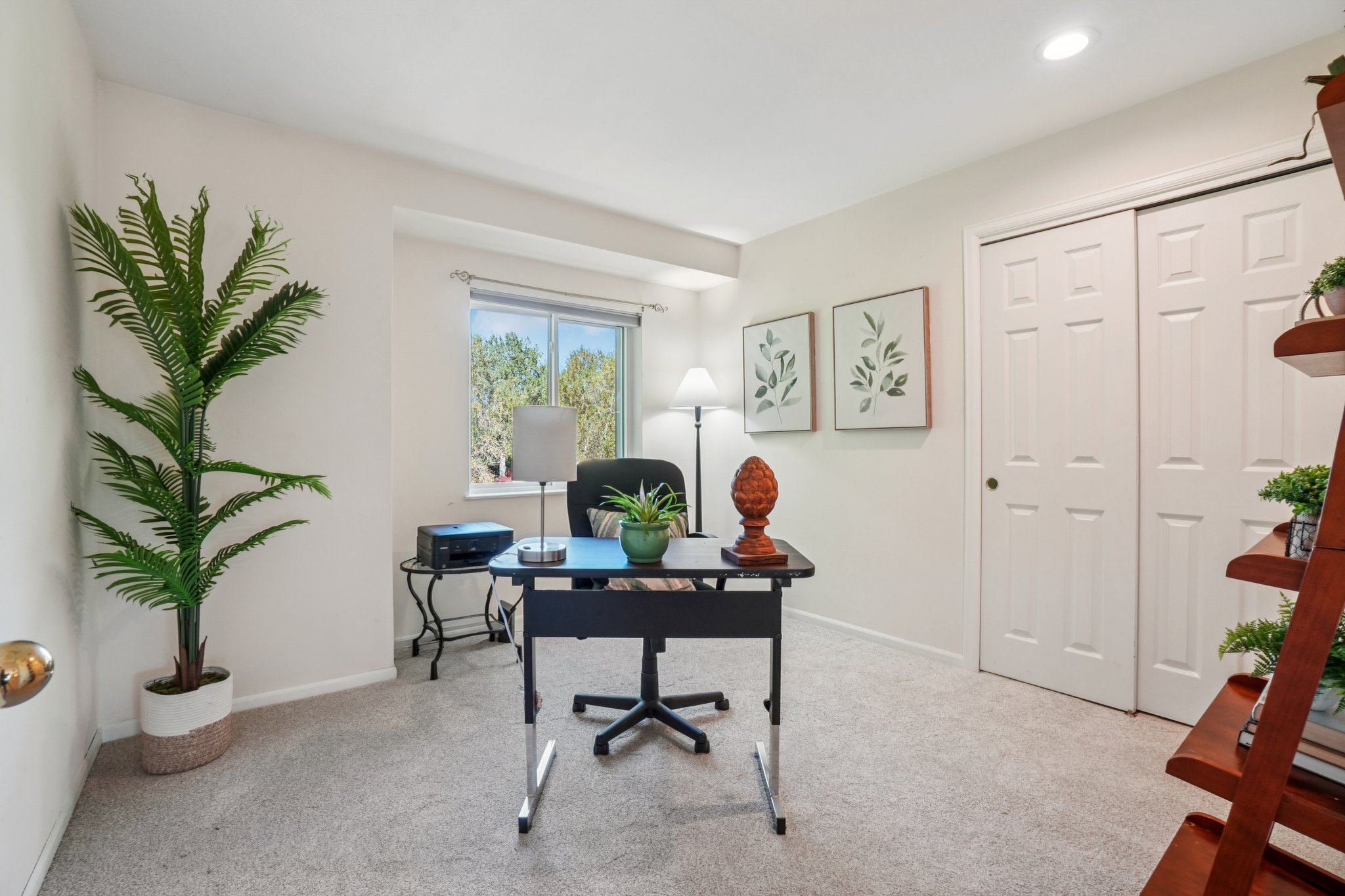 This is an interior shot of a home office. The room features a black desk with a black office chair, a window providing natural light, and neutral-colored walls and carpet. A tall potted plant adds a touch of greenery, and artwork adorns the walls, creating a calm and functional workspace.