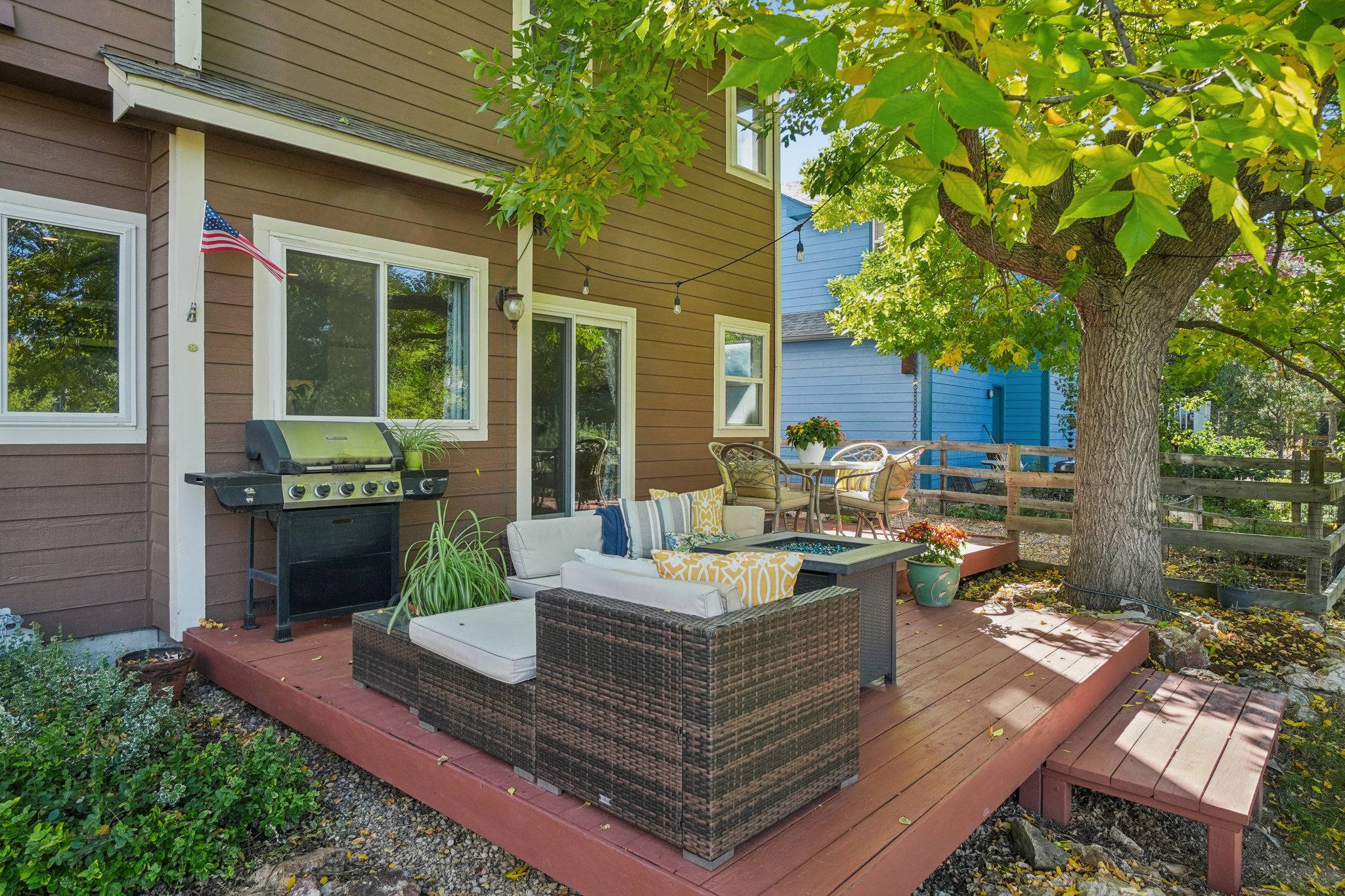 This image showcases a charming outdoor patio area, featuring a brown wicker seating set with comfortable cushions and throw pillows, a fire pit table, and a grill. The patio is constructed of wood and is surrounded by lush greenery and a mature tree, creating a cozy and inviting atmosphere. The exterior of the house is brown with white trim, and an American flag is visible.