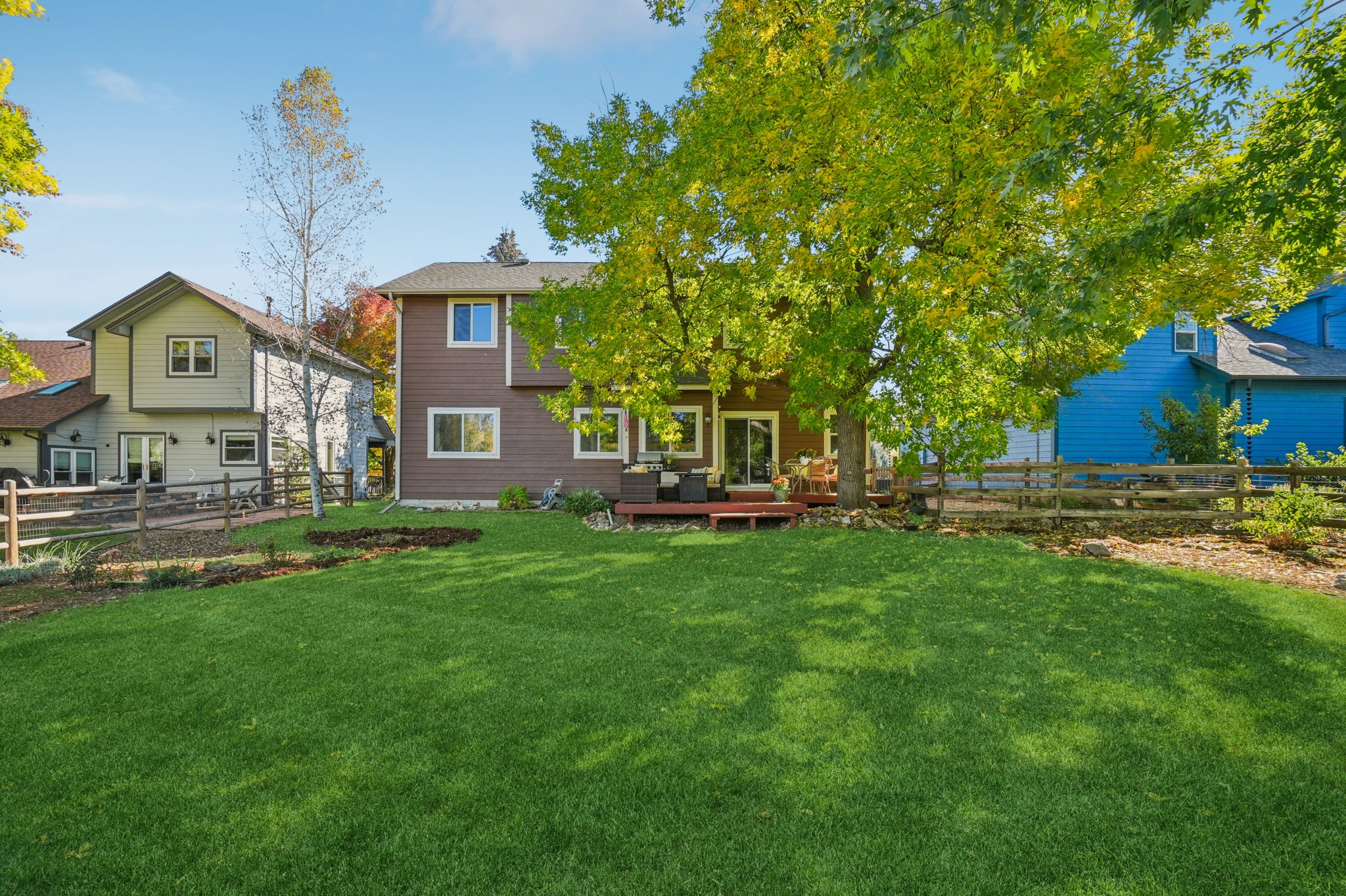 This image showcases a spacious backyard with a lush green lawn, a wooden deck, and mature trees providing shade. The yard is enclosed by a rustic wooden fence, adding to the property's charm. The view includes the rear of the house, featuring brown siding and windows, as well as neighboring houses, creating a sense of community.