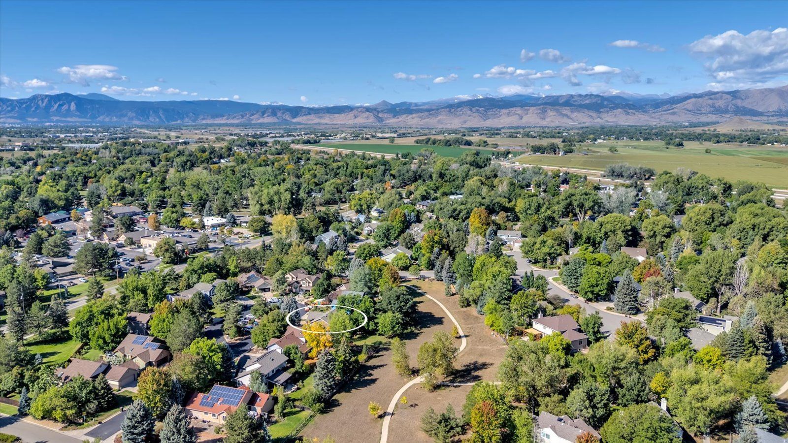 This aerial shot showcases a residential area with lush green trees and well-maintained homes. The property in the center is highlighted with a white circle, surrounded by mature trees and a walking path. In the background, a scenic mountain range adds to the overall appeal of the location.